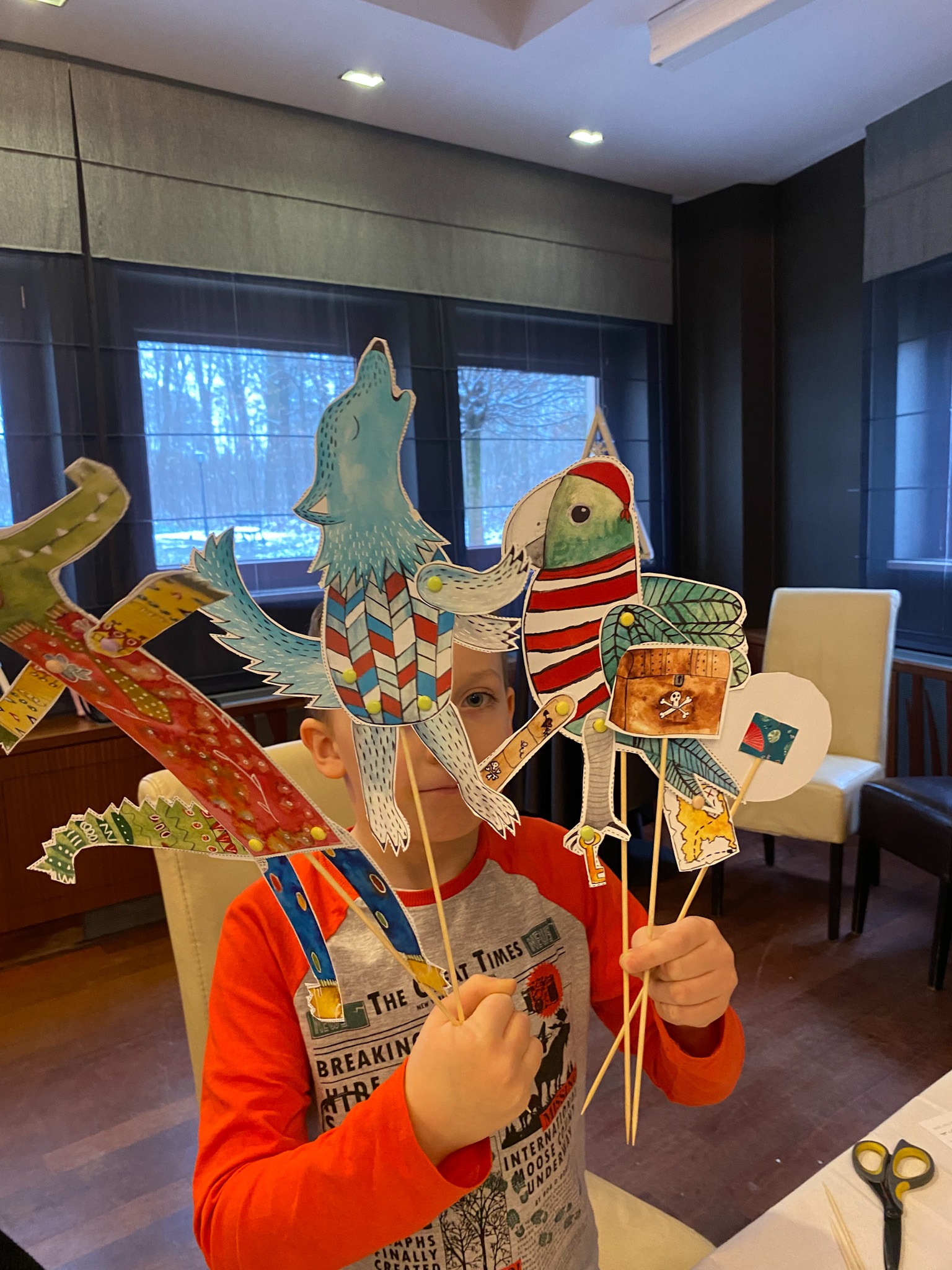 Chłopiec trzymający kukiełki zwierząt na patykach, w tym wilka wyjącego do księżyca, krokodyla i papugę z piracką skrzynią, przygotowane do przedstawienia teatralnego w pomieszczeniu z oknem.