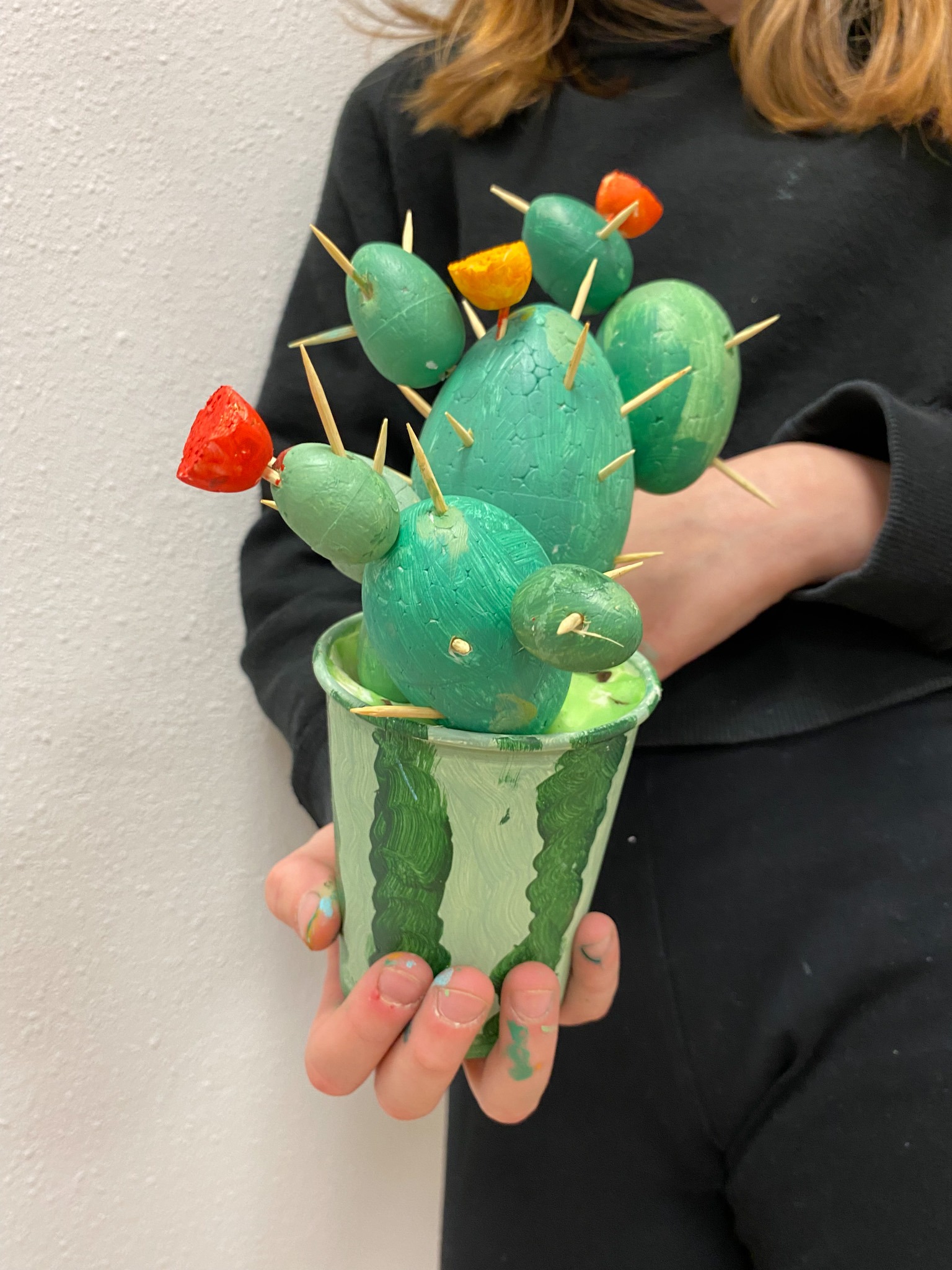 Ręcznie robiony kaktus z pomalowanych jajek i wykałaczek w doniczce z motywem arbuza, trzymany przez osobę z plamami farby na dłoniach.