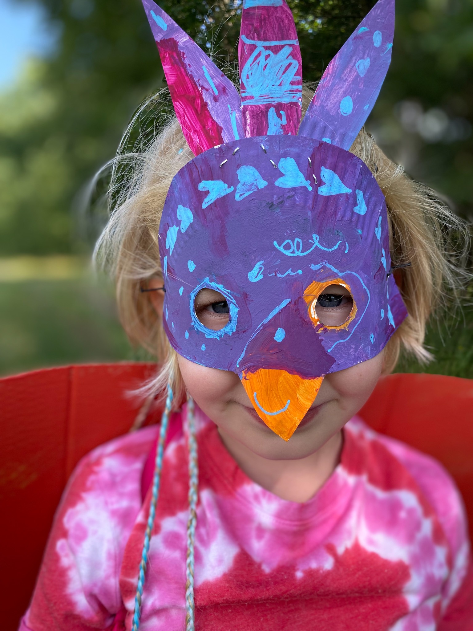 Dziewczynka w ręcznie robionej, pomalowanej masce ptaka z piórami, siedząca na czerwonym krześle na tle zieleni.