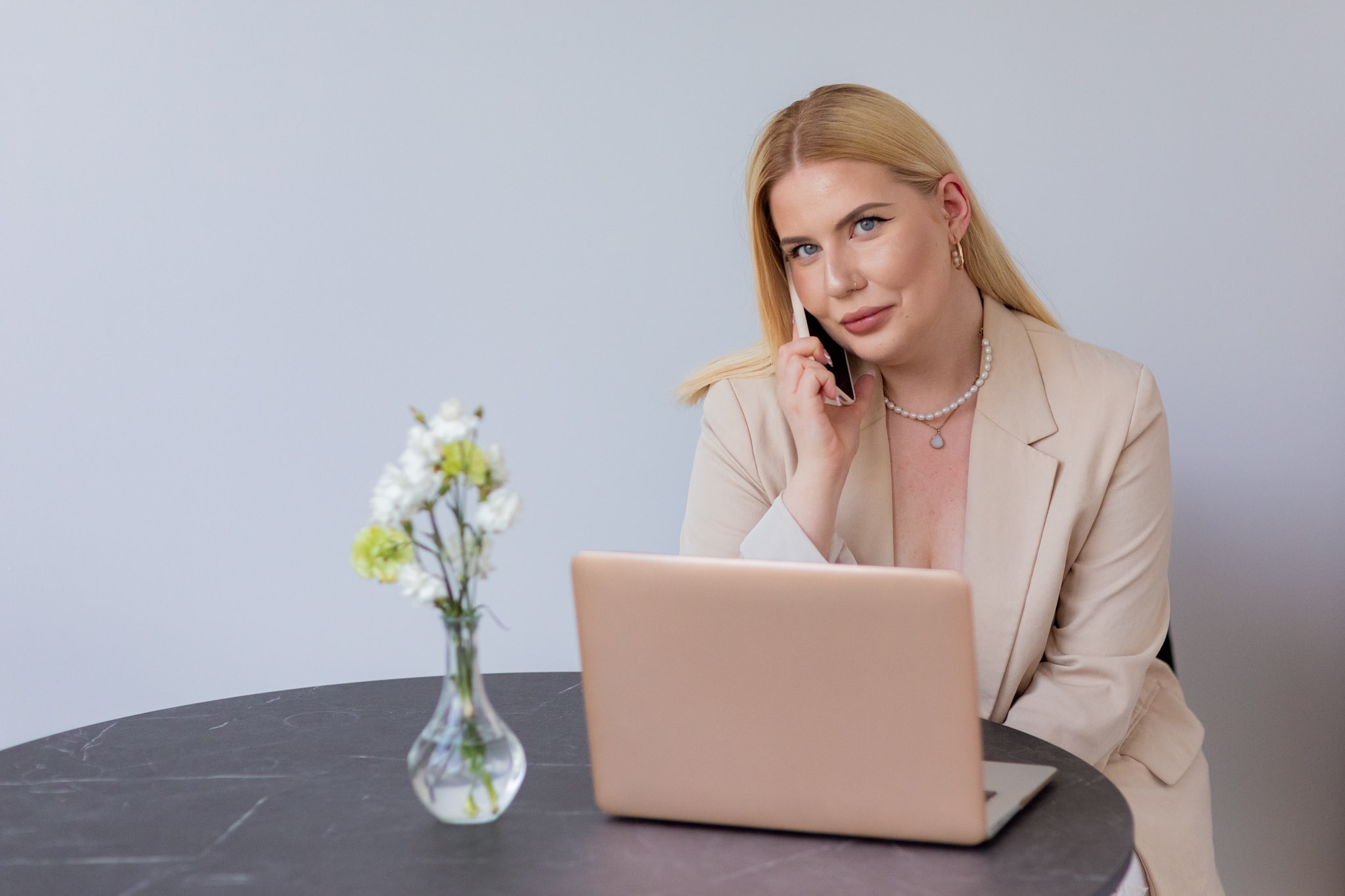 Kobieta w beżowym żakiecie rozmawia przez telefon siedząc przy okrągłym stole z laptopem i wazonem z białymi kwiatami.