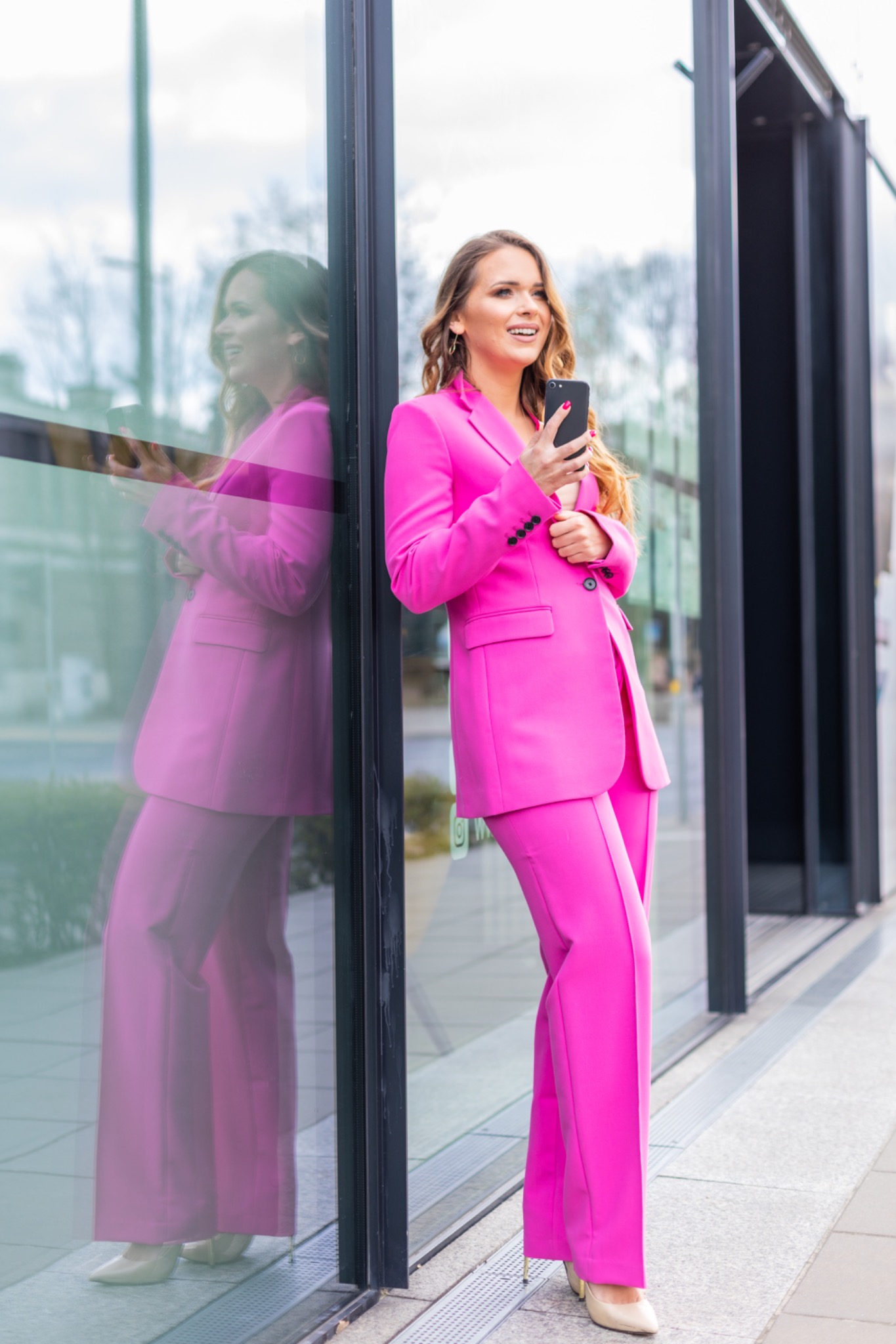 Uśmiechnięta kobieta w różowym garniturze, oparta o przeszkloną ścianę biurowca, trzyma telefon w dłoni.