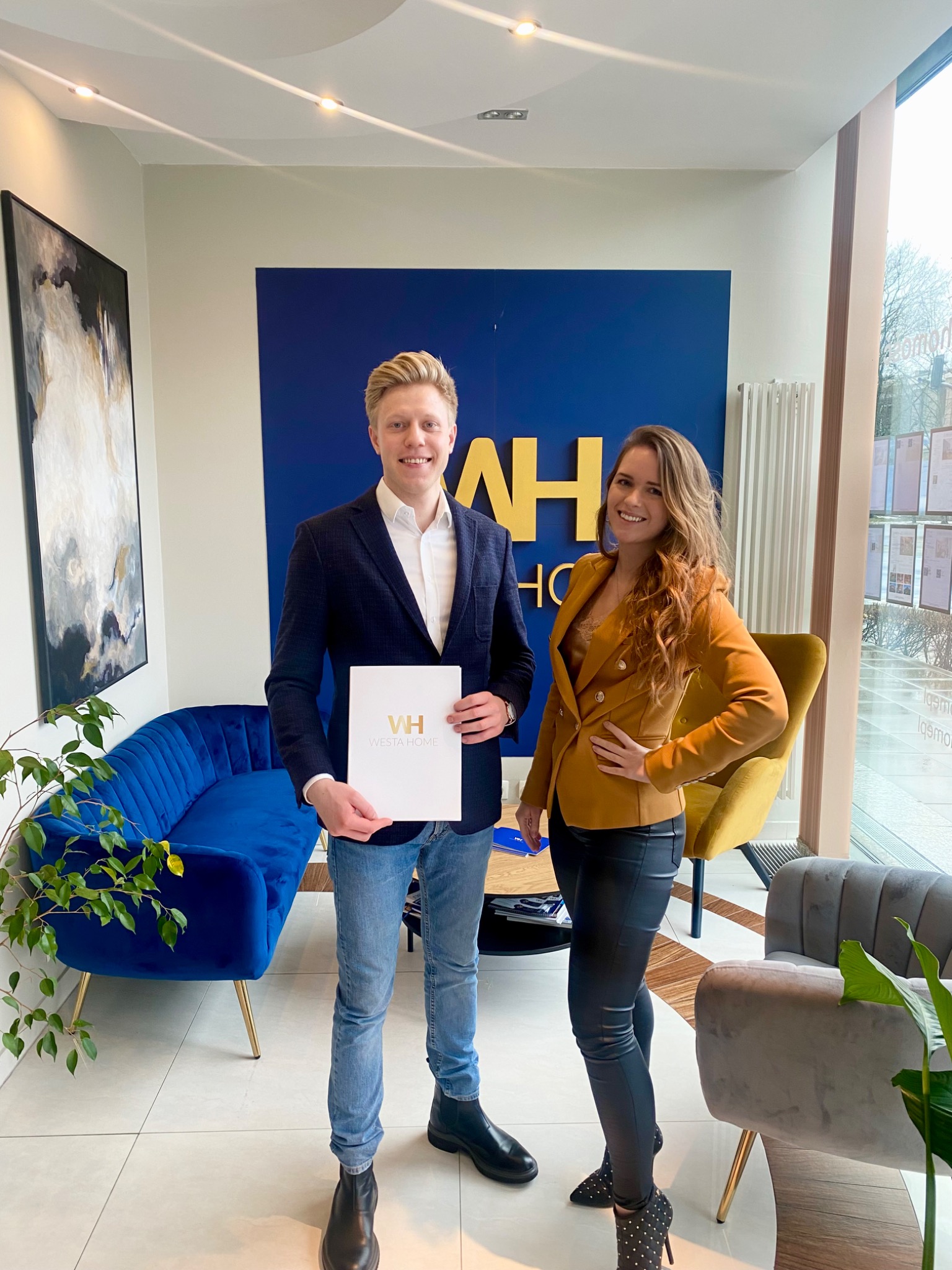 Dwoje uśmiechniętych agentów nieruchomości prezentuje folder firmy Westa Home w nowocześnie urządzonym biurze z niebieską sofą i abstrakcyjnym obrazem.
