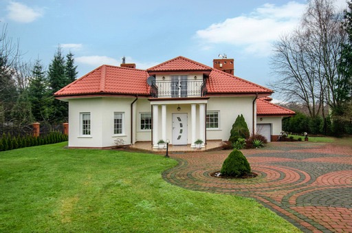 Elegancki dom jednorodzinny z czerwoną dachówką, kolumnami przy wejściu i ozdobnym brukowanym podjazdem w kształcie wachlarza.