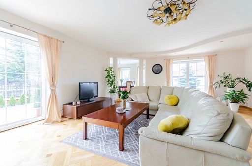 Jasny salon z narożną sofą, drewnianym stolikiem kawowym na wzorzystym dywanie, telewizorem na niskiej komodzie, dużymi oknami z zasłonami i dekoracyjnym żyrandolem.