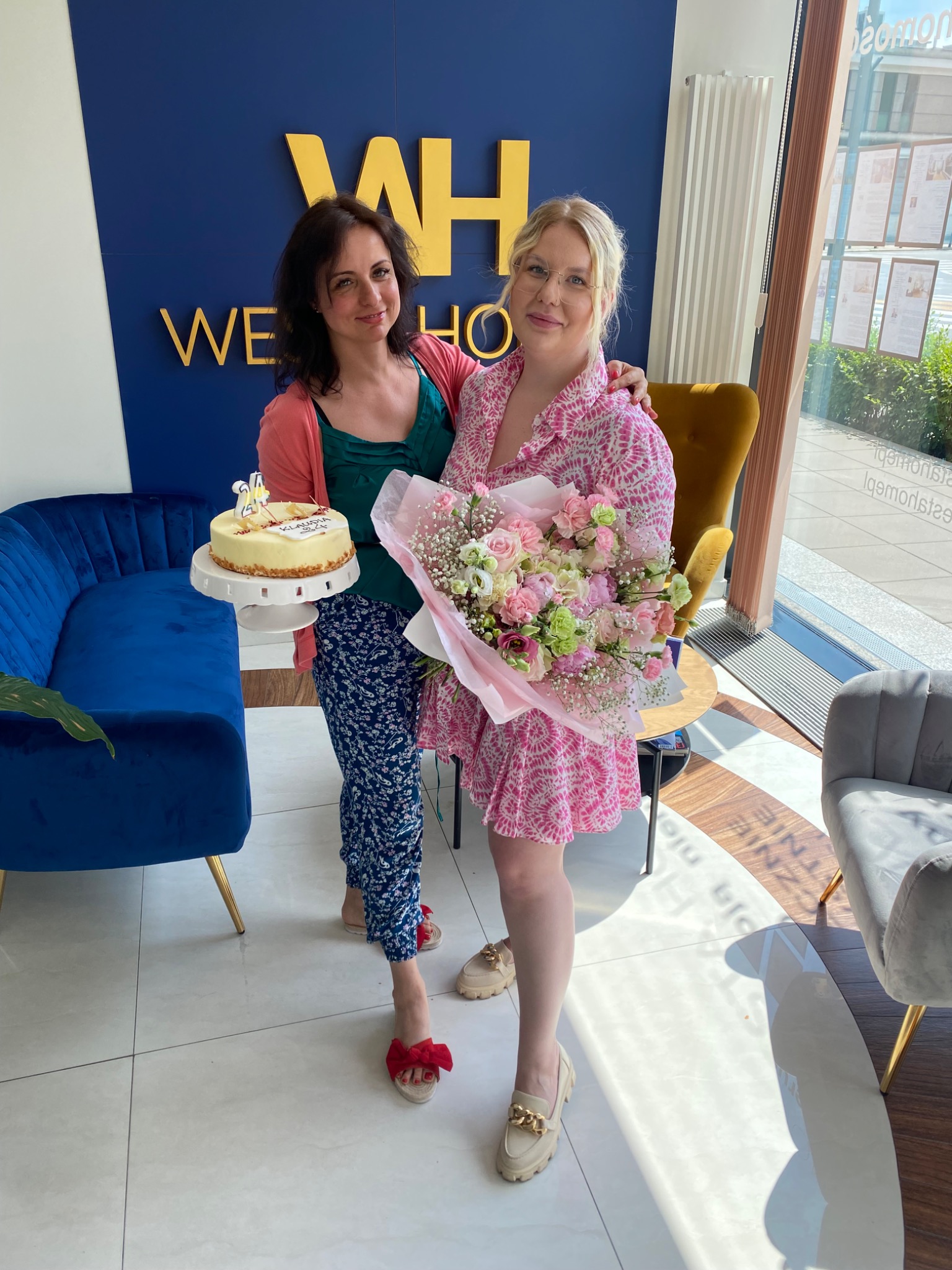 Dwie uśmiechnięte kobiety w biurze nieruchomości 'WH We Home' świętują z tortem urodzinowym i bukietem kwiatów na tle niebieskiej ściany z logo i okna z ofertami.