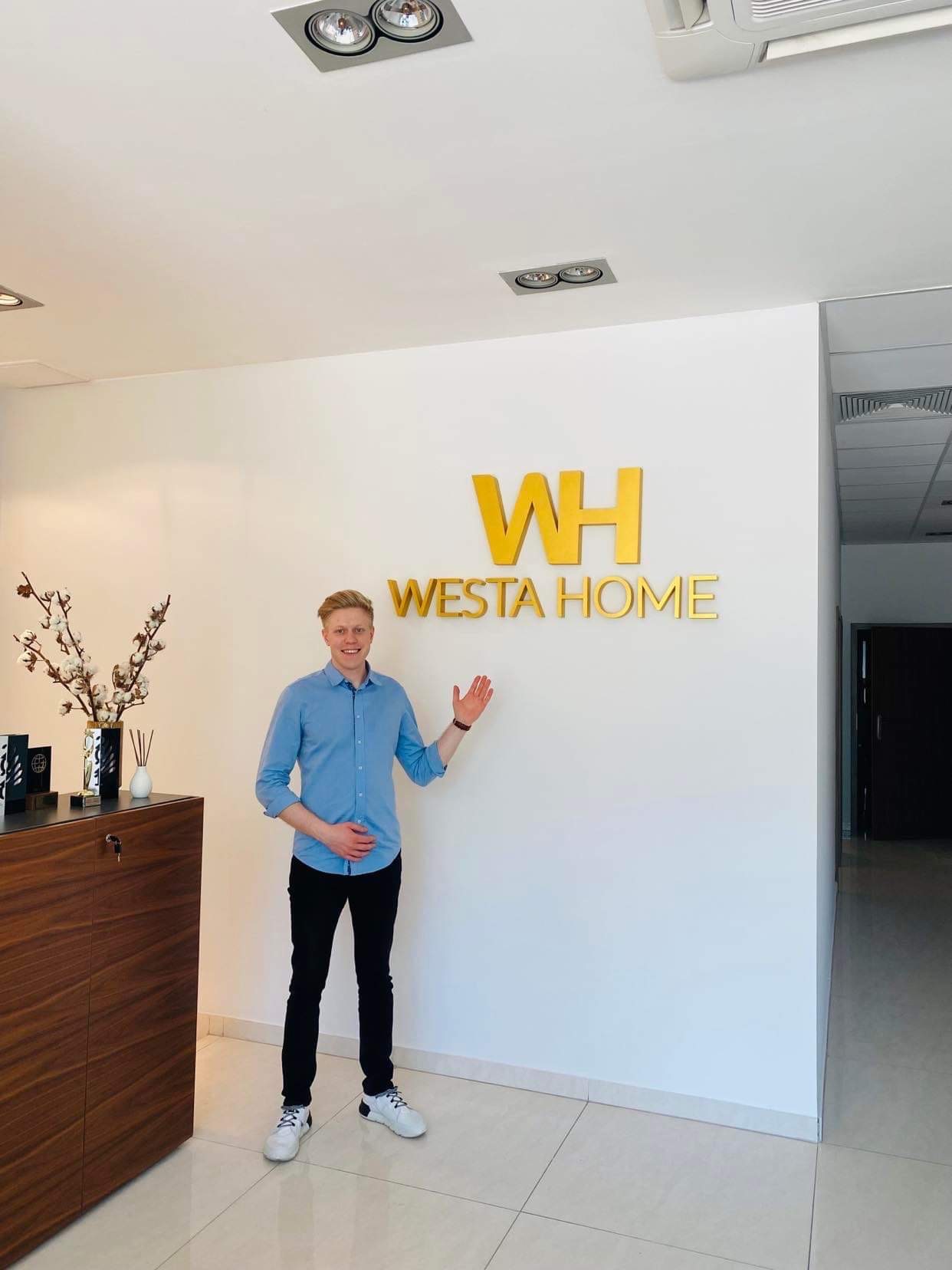 Pracownik biura 'WESTA HOME' w Warszawie prezentuje logo firmy na białej ścianie. W tle widoczny minimalistyczny wystrój wnętrza, w tym drewniana szafka i dekoracyjne gałązki bawełny.