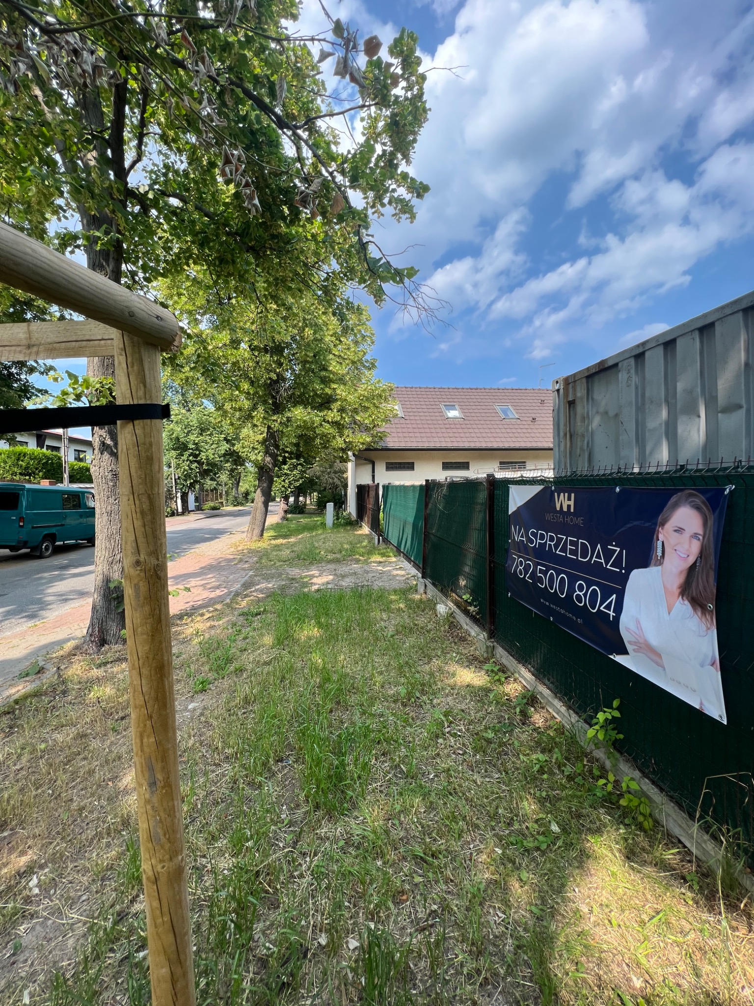 Baner reklamowy 'Na Sprzedaż!' z wizerunkiem kobiety, umieszczony na zielonym ogrodzeniu siatkowym wzdłuż chodnika z trawą, w tle dom z czerwonym dachem i niebieski samochód dostawczy