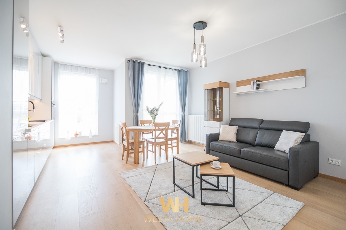 Jasne wnętrze mieszkania z drewnianą podłogą, szarą sofą, drewnianym stołem z krzesłami, nowoczesnym oświetleniem i dywanem w geometryczne wzory. Widoczna również biała zabudowa kuchenna...