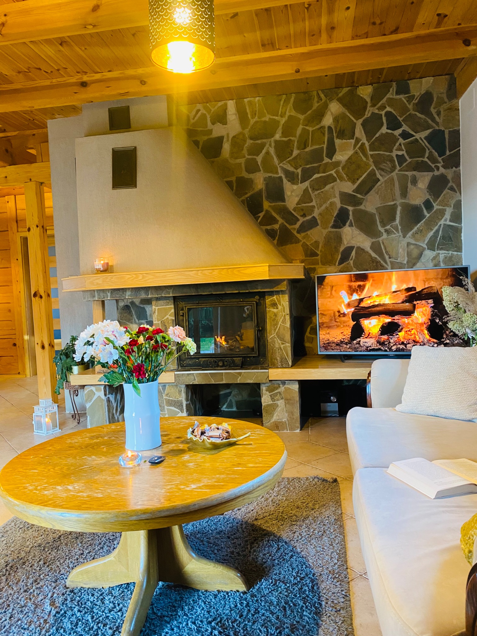 Przytulny salon z kamiennym kominkiem, płonącym drewnem na ekranie telewizora, okrągłym drewnianym stolikiem z kwiatami i słodyczami, oraz otwartą książką na białej sofie.