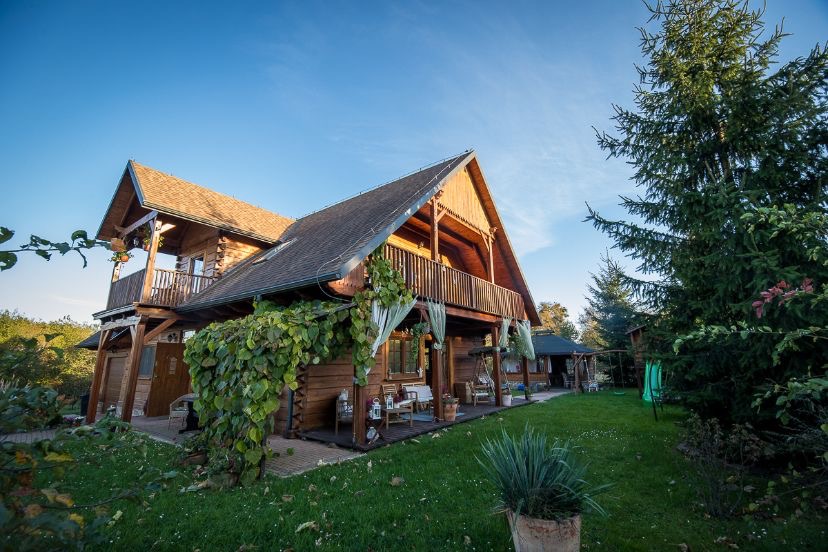 Drewniany dom z dwoma balkonami, porośnięty bluszczem, z widocznym ogrodem i świerkiem na tle błękitnego nieba.