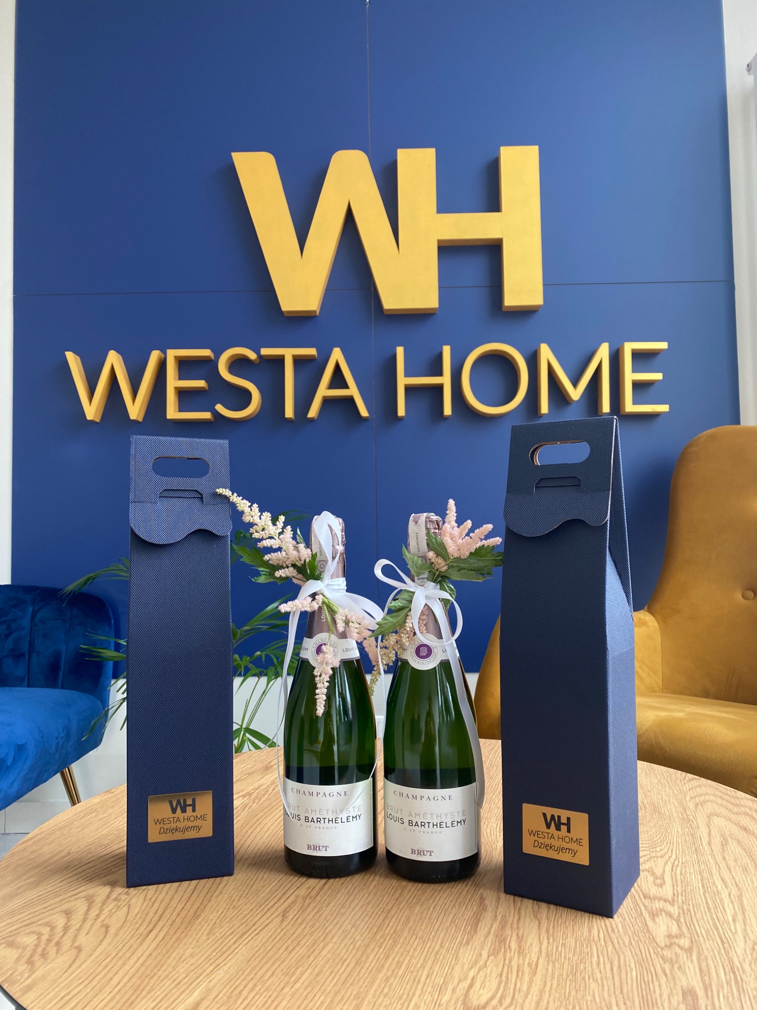 Dwie butelki szampana Louis Barthelemy udekorowane kwiatami, umieszczone obok granatowych torebek prezentowych z logo Westa Home na drewnianym stoliku, z niebieską ścianą i złotym napisem Westa...
