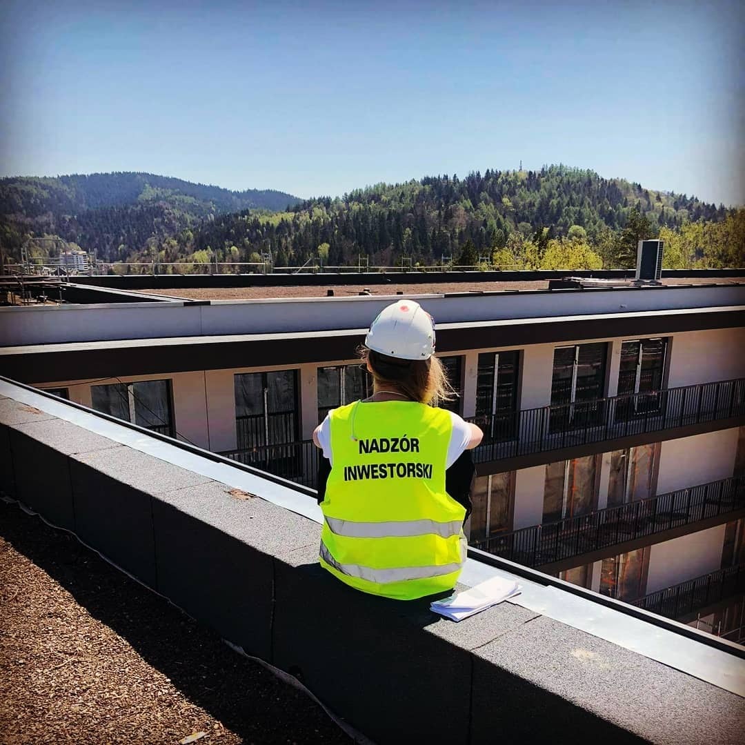 Osoba w kasku i żółtej kamizelce z napisem 'Nadzór Inwestorski' siedzi na krawędzi dachu budynku wielorodzinnego, w tle zalesione wzgórza i błękitne niebo.