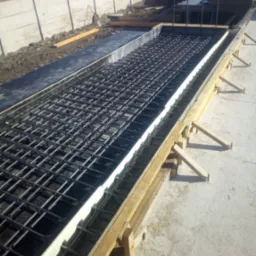 Przygotowanie zbrojenia w szalunku z drewnianymi podporami i izolacją termiczną na budowie. Widoczne stalowe pręty zbrojeniowe ułożone w siatkę.