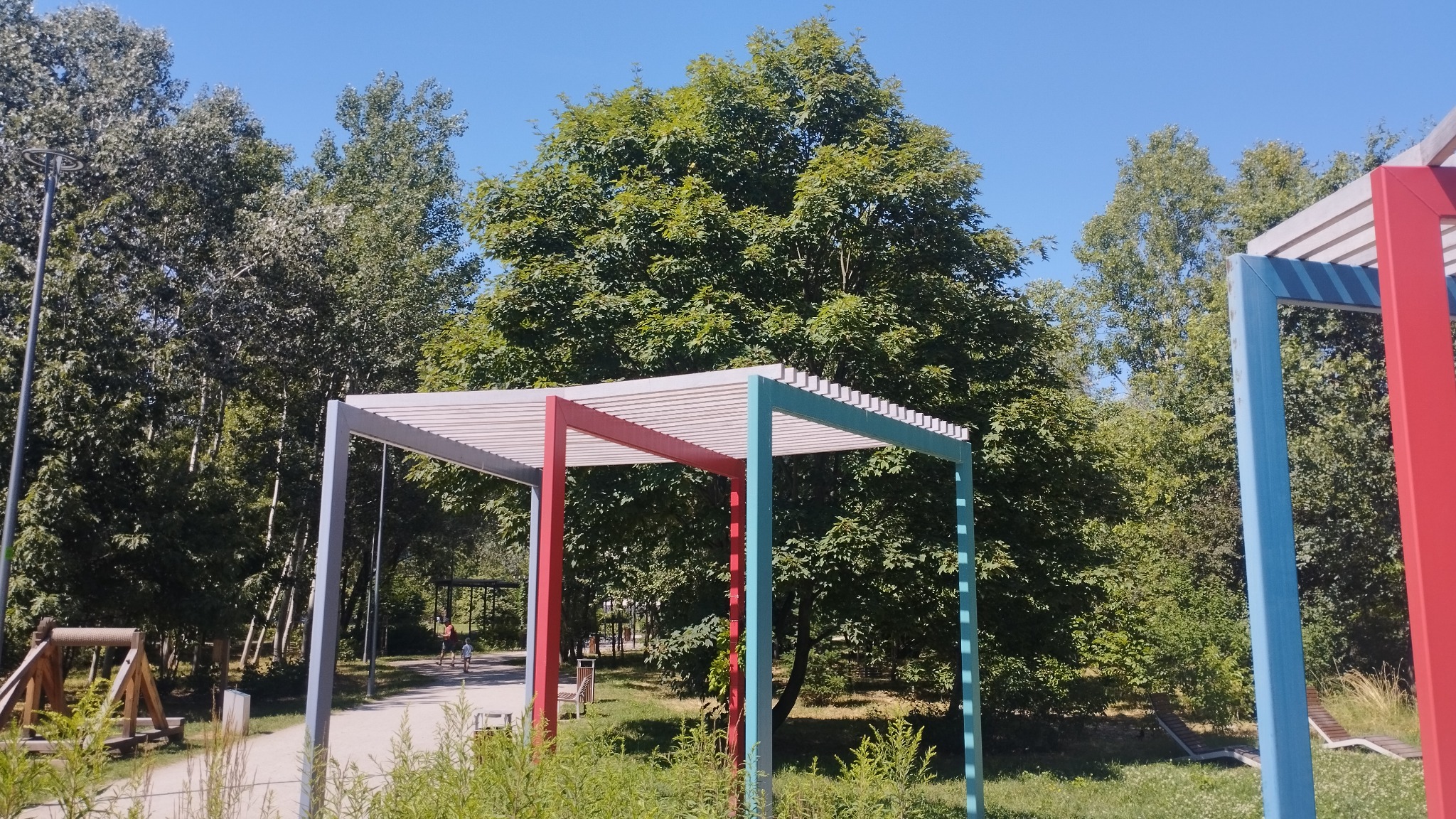 Kolorowa pergola z elementami czerwonymi, niebieskimi i szarymi w parku miejskim, na tle zieleni drzew i błękitnego nieba.