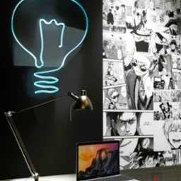 Biurko z laptopem, lampką i neonową lampą w kształcie żarówki na tle ściany pokrytej mangą.