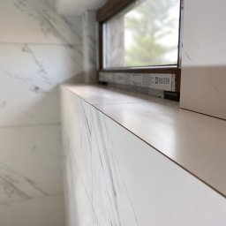 Yurii Yatsyshyn Yat-Fliz - Ściana i parapet wyłożone białymi płytkami imitującymi marmur, z widocznym oknem z brązową ramą i folią ochronną producenta na profilu.