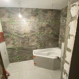 Yurii Yatsyshyn Yat-Fliz - Łazienka w trakcie układania płytek imitujących marmur na ścianach, z użyciem poziomych sznurków i zielonych klipsów dystansowych, widoczna biała wanna narożna z obudową.