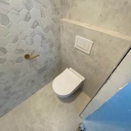 Yurii Yatsyshyn Yat-Fliz - Wykończona łazienka z toaletą podwieszaną, heksagonalnymi płytkami imitującymi marmur na ścianie, złotym uchwytem na papier toaletowy i betonową ścianą z przyciskiem spłukującym.