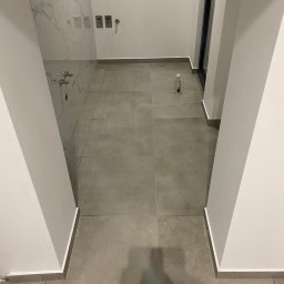 Yurii Yatsyshyn Yat-Fliz - Szare płytki podłogowe w łazience z marmurową ścianą i butelką środka czyszczącego w rogu.