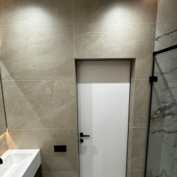 Yurii Yatsyshyn Yat-Fliz - Nowoczesna łazienka z beżowymi płytkami imitującymi kamień, białą umywalką z szafką z drewnianym frontem i białą, minimalistyczną, ukrytą w ścianie, wewnętrzną ościeżnicą z czarną klamką.