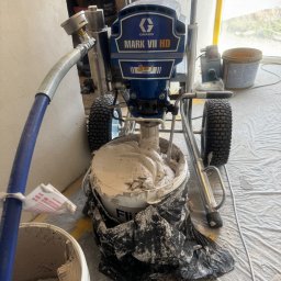 Yurii Yatsyshyn Yat-Fliz - Agregat malarski Graco Mark VII HD z mieszadłem w wiadrze z farbą, przygotowany do pracy w Warszawie. Podłoże zabezpieczone folią malarską.