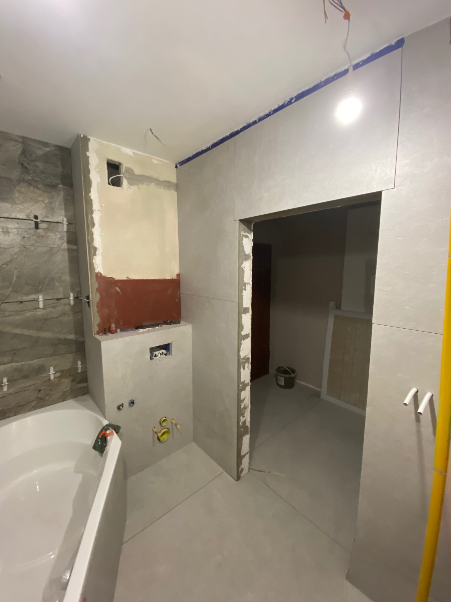 Łazienka w trakcie remontu: widoczna wanna, częściowo wykończona ściana z szarymi płytkami imitującymi kamień, fragment ściany przygotowany pod glazurę z widocznymi instalacjami...