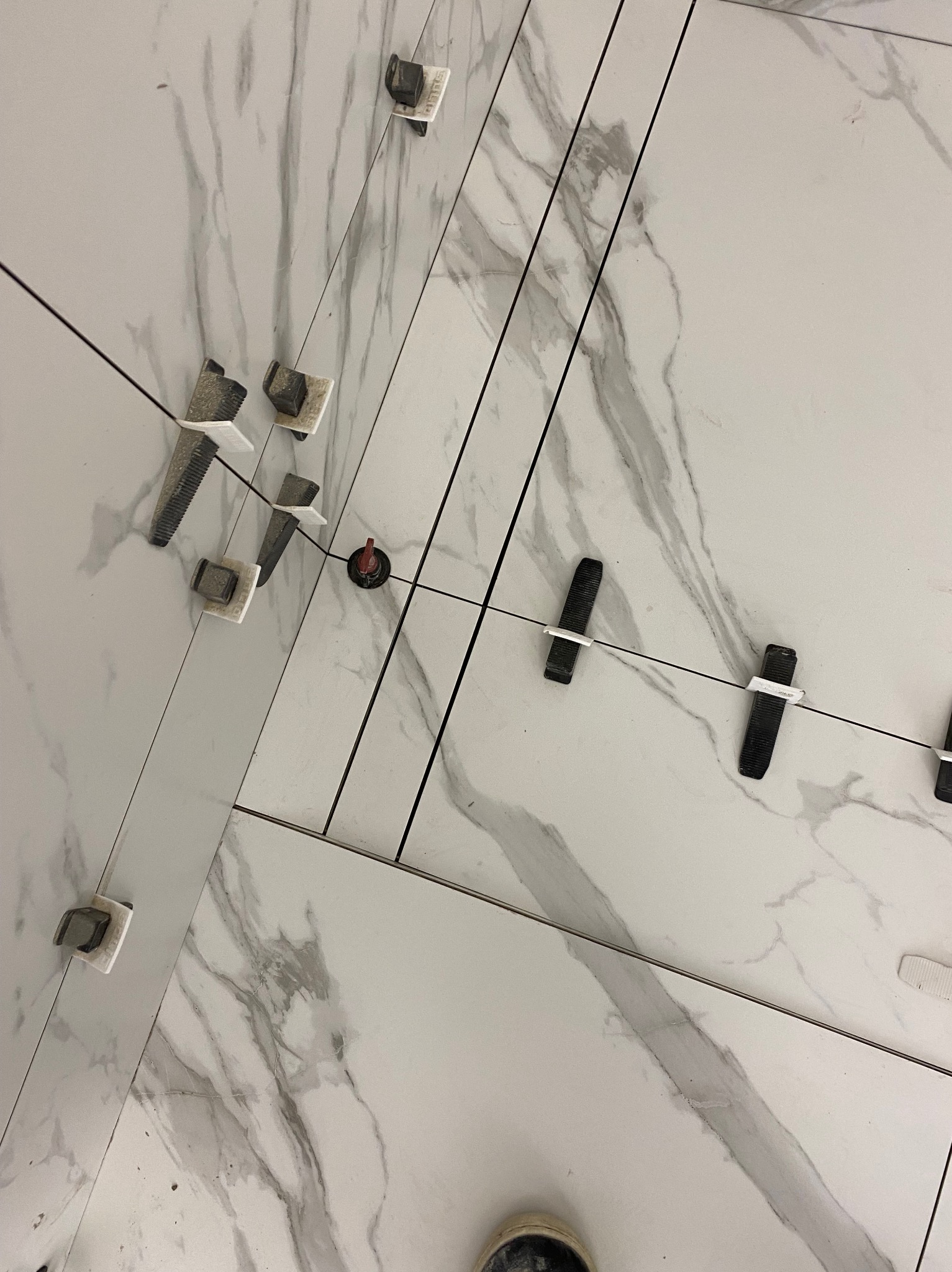 Układanie białych płytek imitujących marmur z użyciem systemu poziomowania płytek; widoczne klipsy, kliny i zaciskarka.