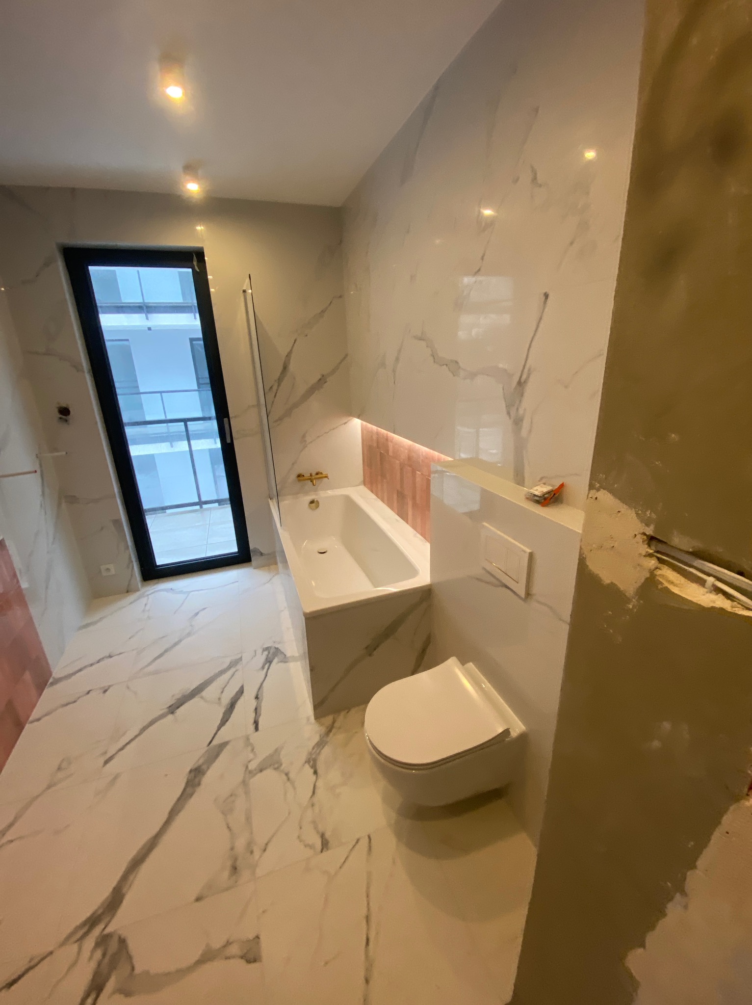 Nowoczesna łazienka w trakcie wykańczania: białe marmurowe płytki, wanna obudowana płytkami z akcentem różowego, wisząca toaleta i fragment ściany w stanie surowym.