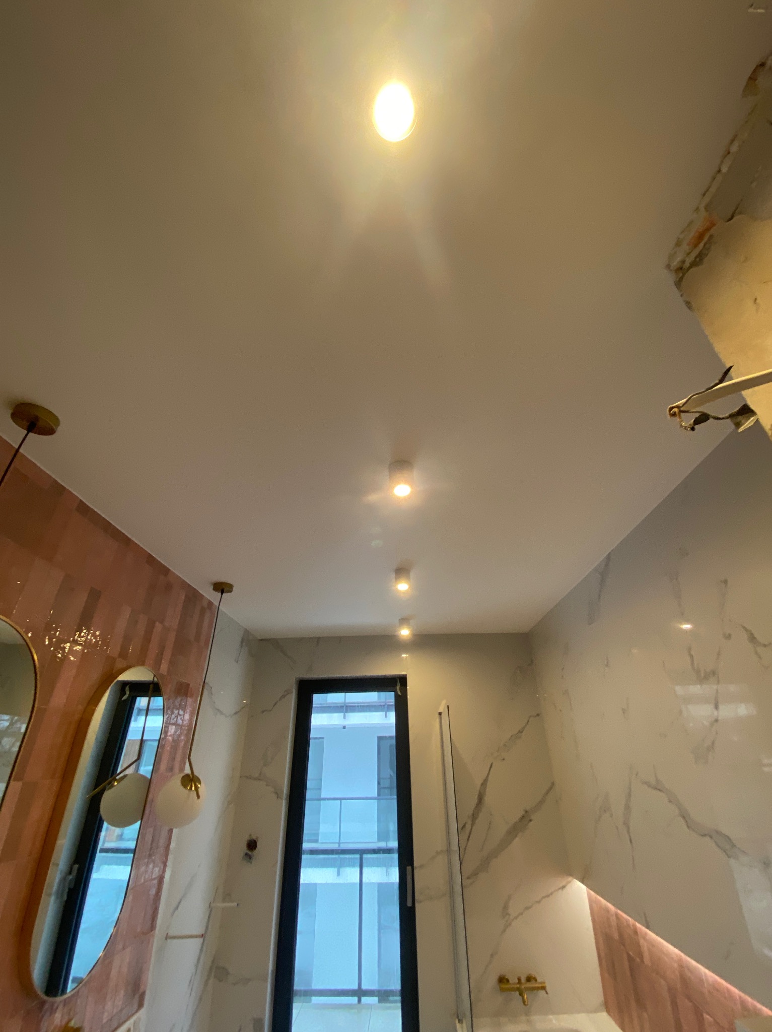 Wnętrze łazienki w trakcie prac wykończeniowych; widoczny fragment niezabezpieczonego narożnika sufitu, różowe płytki na ścianie w połączeniu z marmurowym wzorem, rząd punktowych lamp sufitowych.