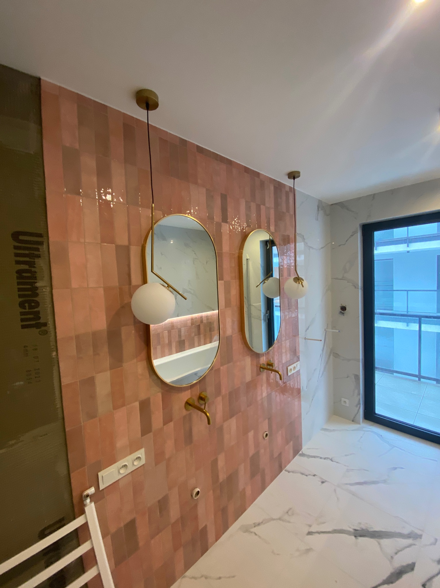 Nowoczesna łazienka w trakcie wykańczania: różowa, prostokątna glazura, złote baterie, owalne lustra z kulistymi lampami, marmurowe płytki podłogowe, widoczna płyta ochronna Ultrament.