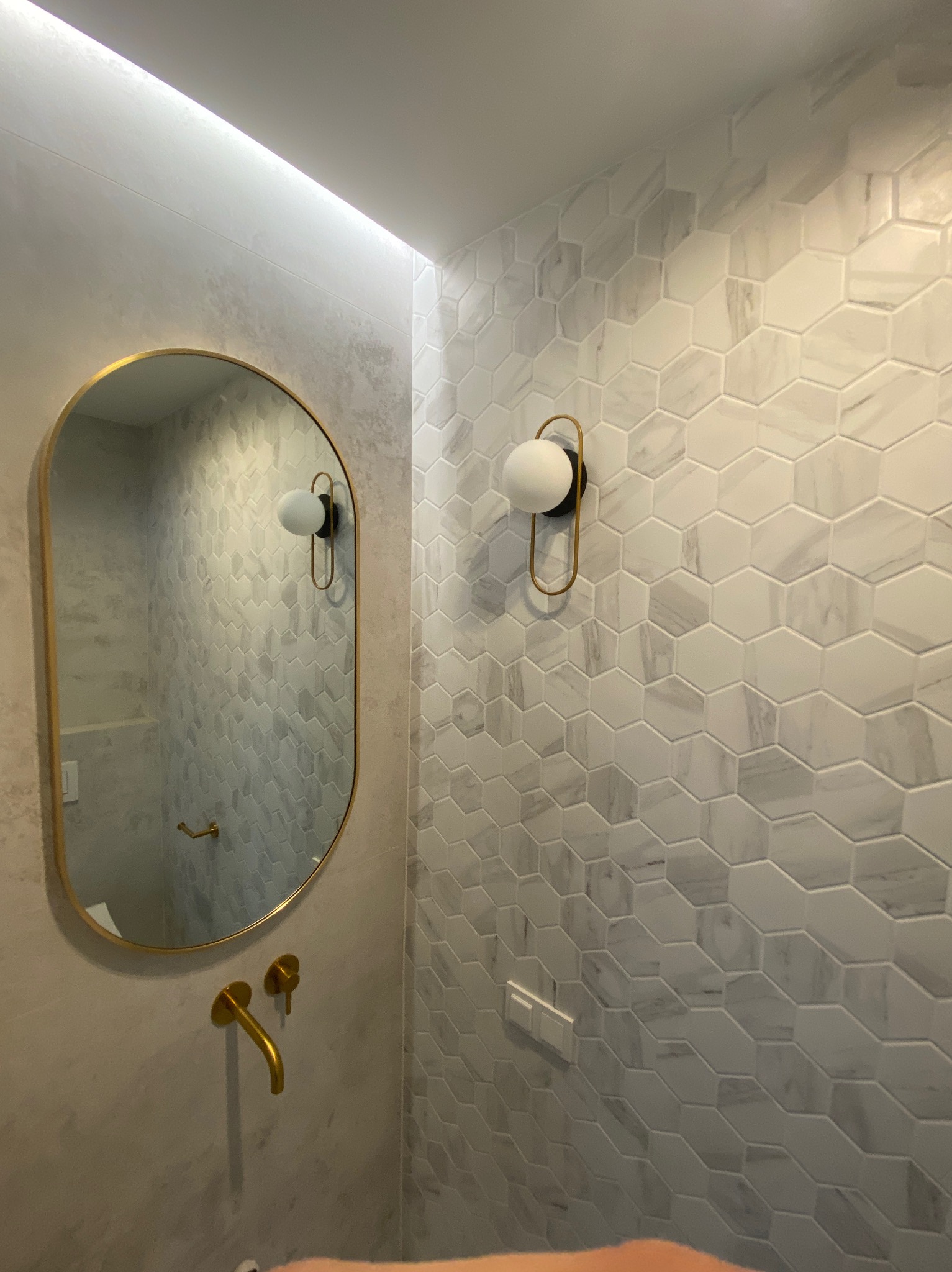 Elegancka łazienka z heksagonalnymi płytkami marmuropodobnymi, owalne lustro w złotej ramie i złota bateria umywalkowa podtynkowa, nowoczesny kinkiet z kulistym kloszem.