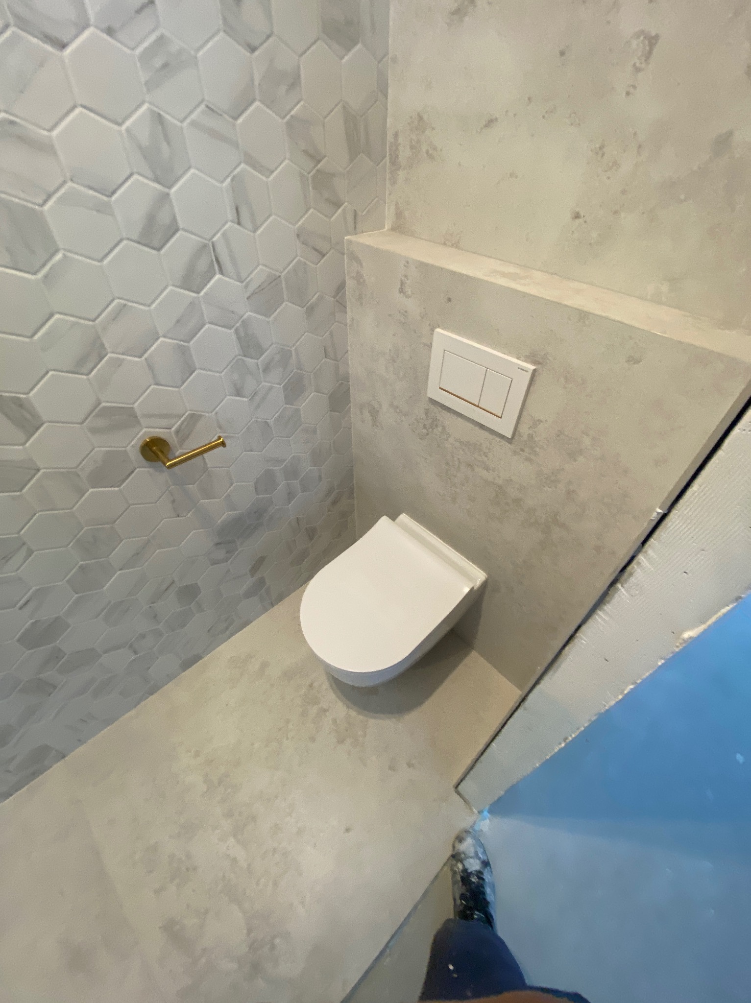 Wykończona łazienka z toaletą podwieszaną, heksagonalnymi płytkami imitującymi marmur na ścianie, złotym uchwytem na papier toaletowy i betonową ścianą z przyciskiem spłukującym.