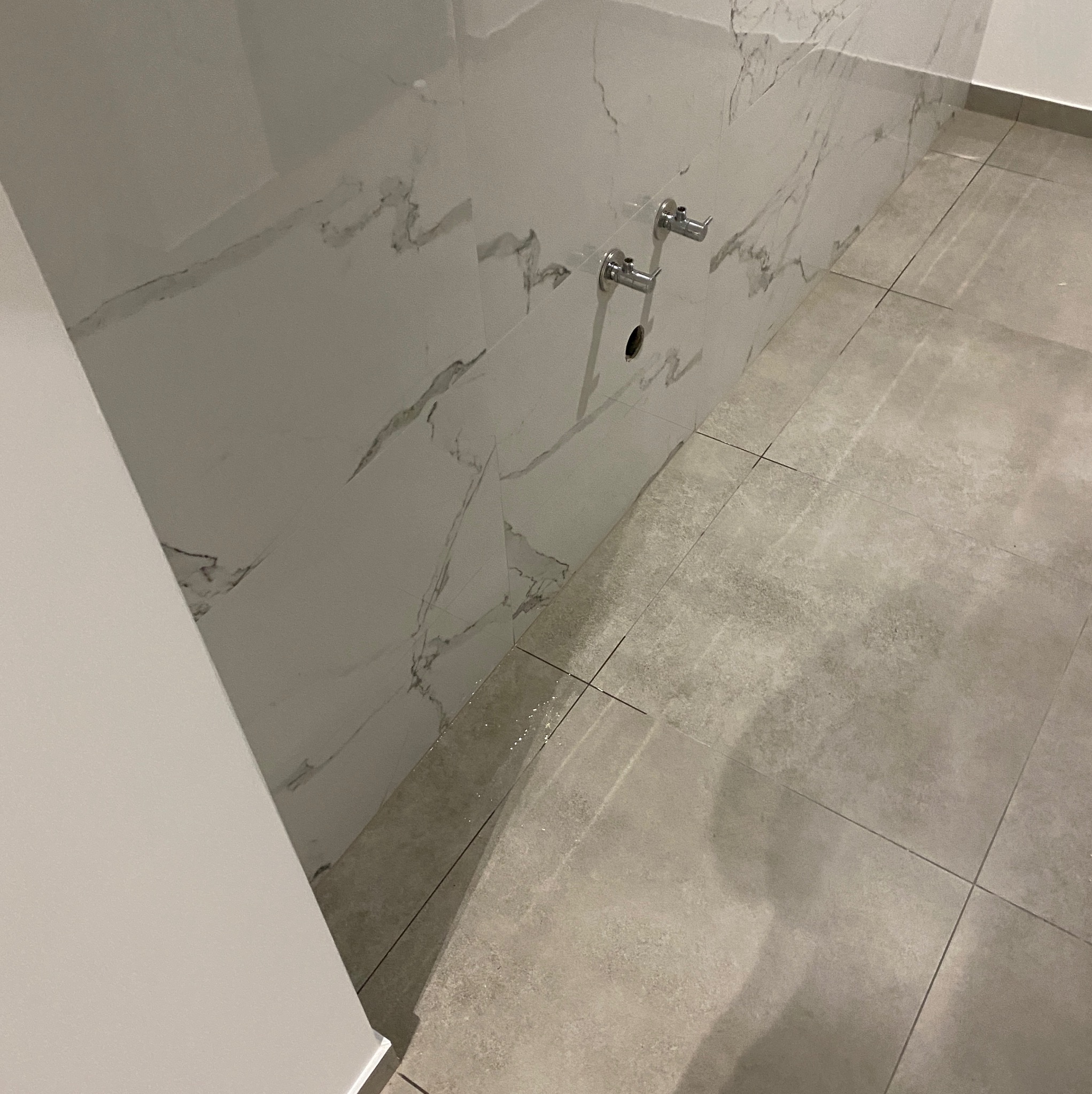 Fragment łazienki w trakcie wykańczania: ściana z płytek imitujących marmur, zamontowane zawory, szare płytki podłogowe, widoczne niedociągnięcia przy łączeniu płytek.