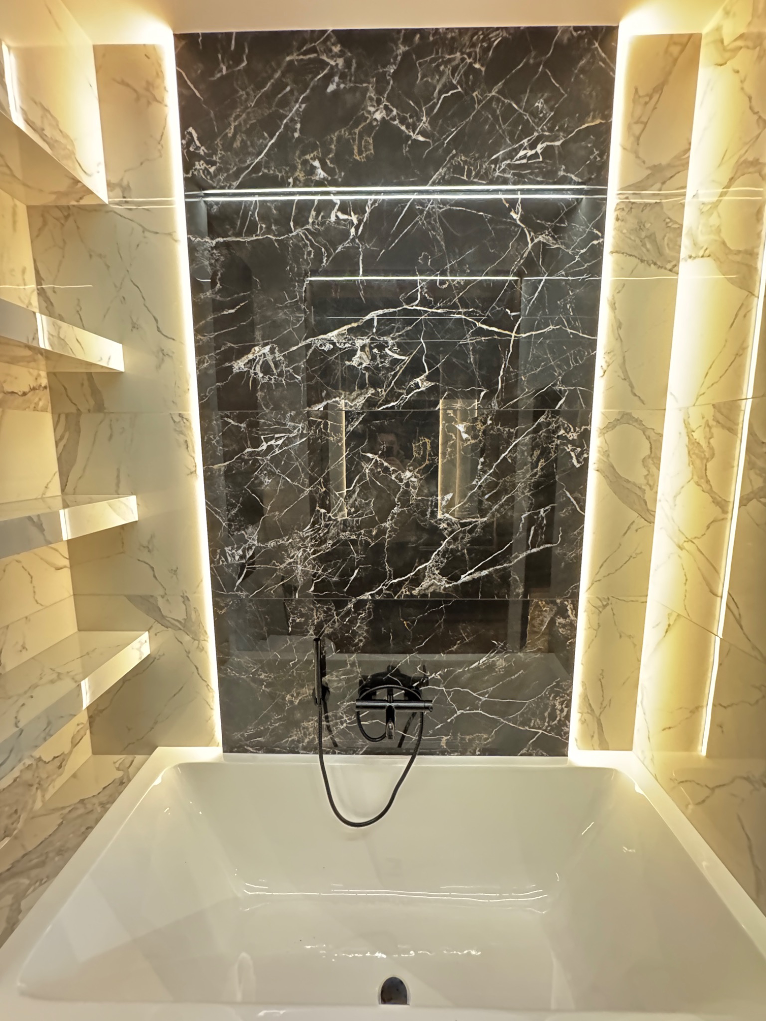 Nowoczesna łazienka z wanną, czarnymi płytkami imitującymi marmur na ścianie z wnęką i podświetlanymi półkami z jasnego marmuru po bokach.