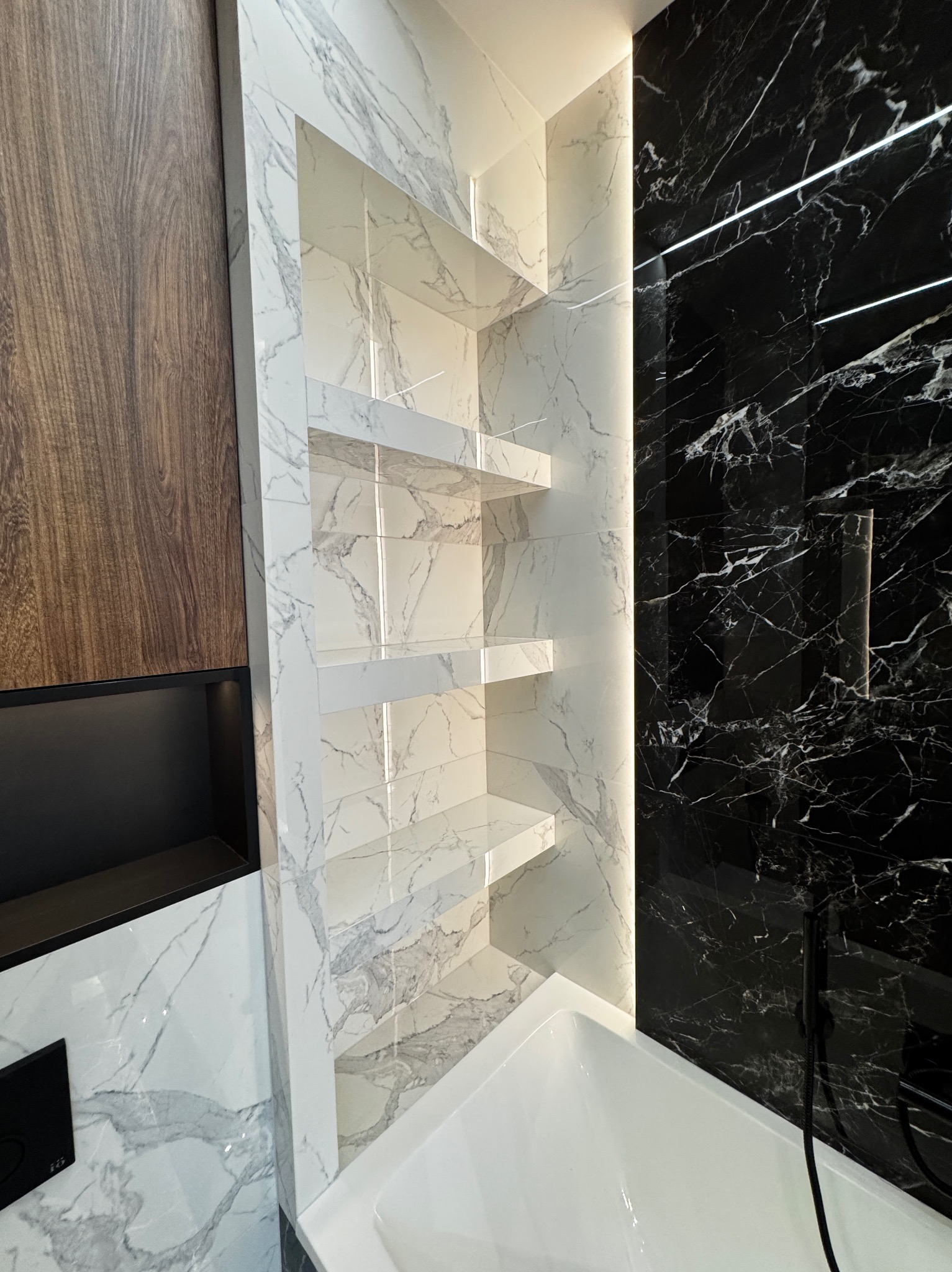 Nowoczesna łazienka z czarno-białymi marmurowymi płytkami, podświetlane półki z białego marmuru, drewniana okładzina i biała wanna.