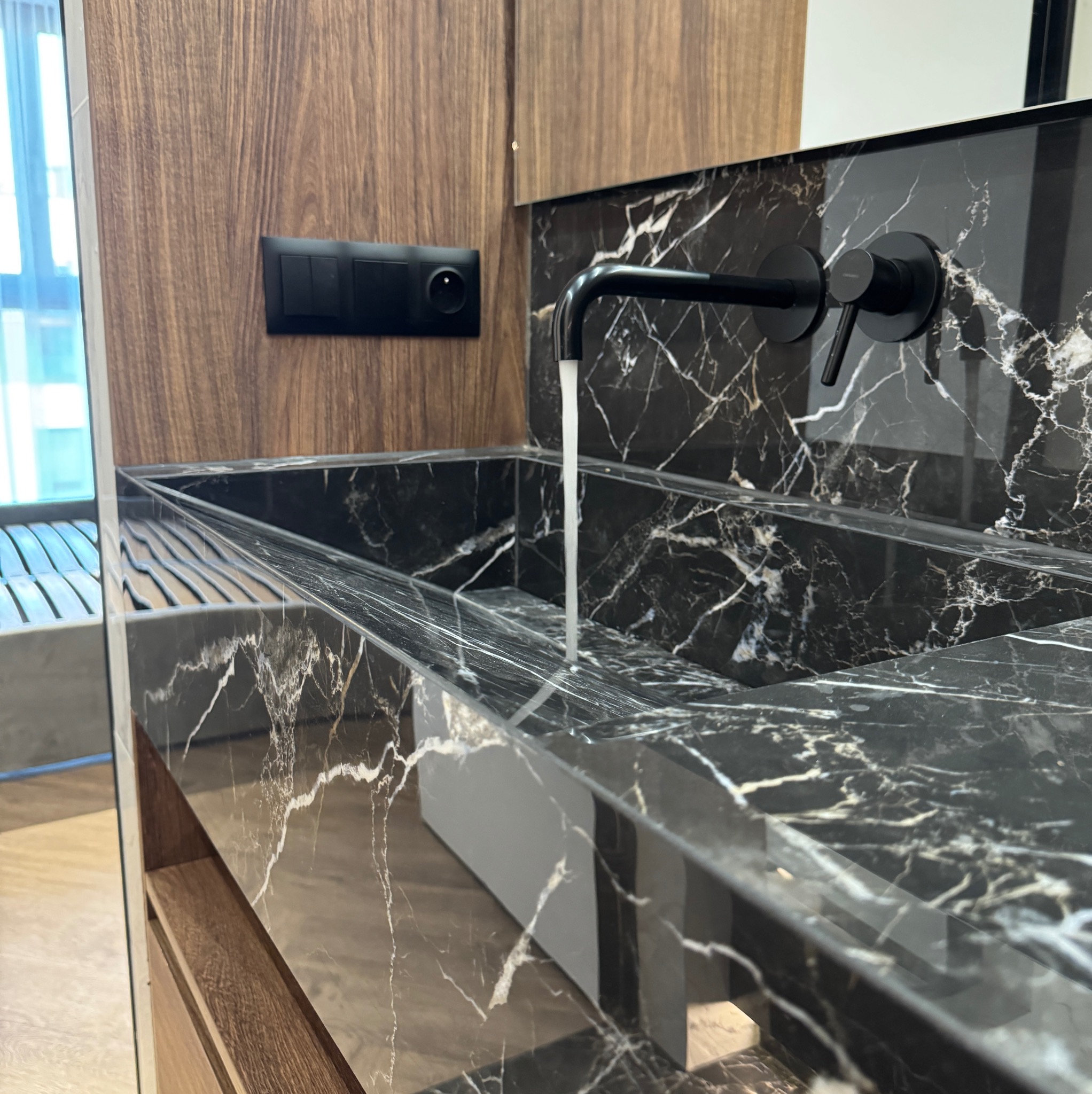 Nowoczesna łazienka: Czarny marmurowy blat umywalki z wpuszczonym zlewem, czarna bateria z wypływającą wodą, drewniana szafka i czarny panel z włącznikami.