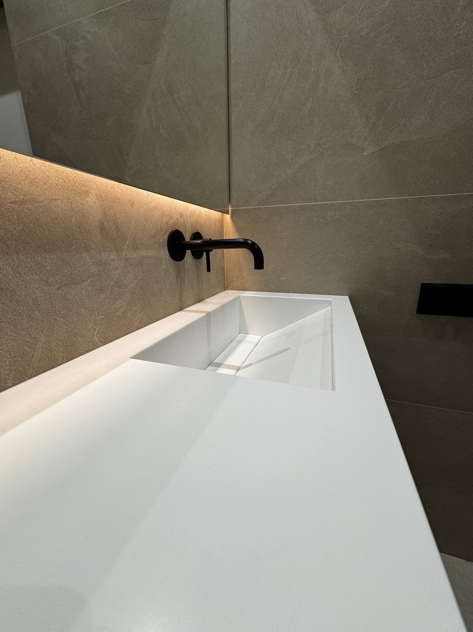 Nowoczesna łazienka z białą, geometryczną umywalką z konglomeratu, czarną, matową armaturą ścienną i podświetlanym lustrem na tle płytek imitujących kamień.