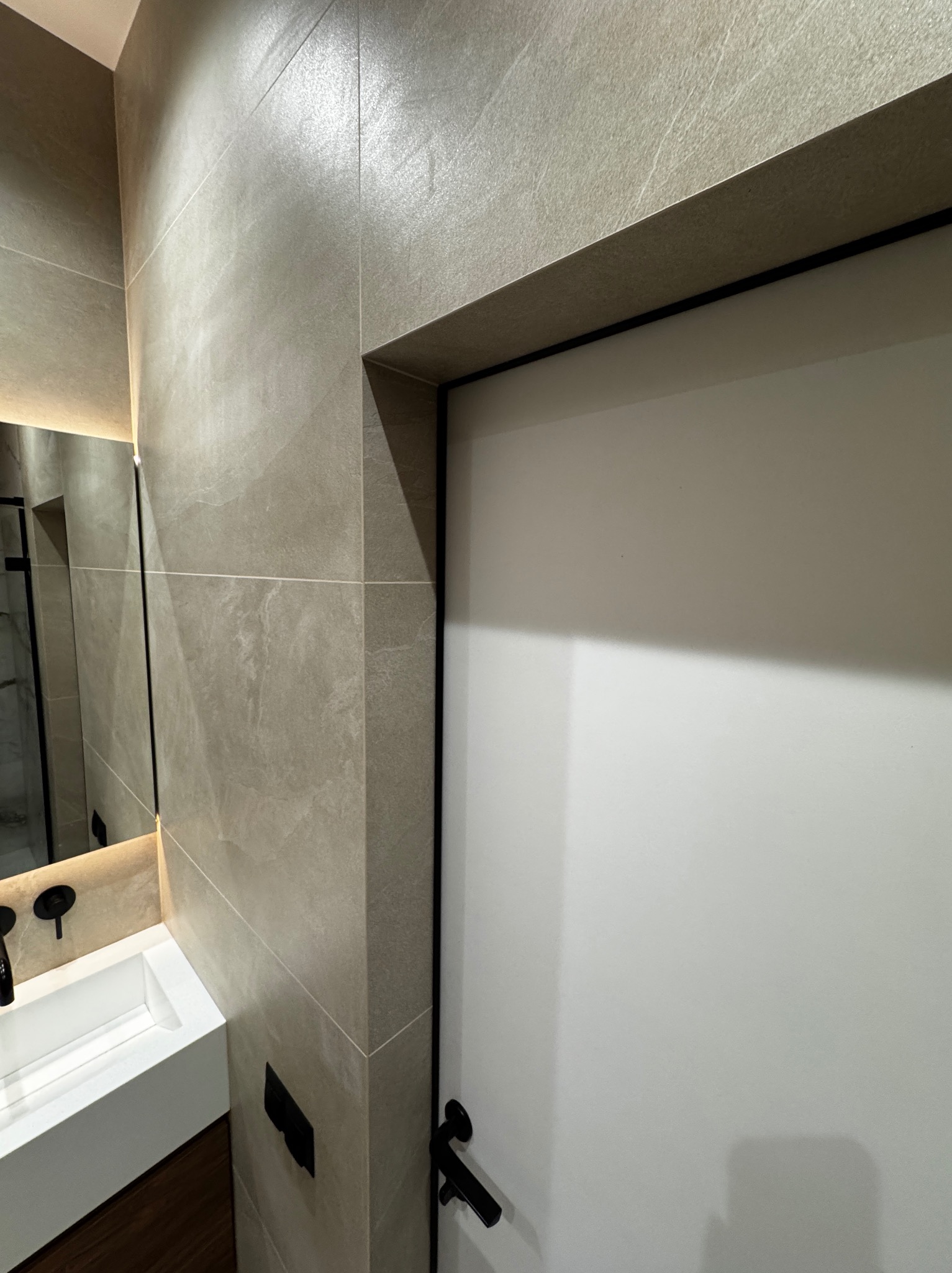 Nowoczesna łazienka z beżowymi płytkami imitującymi kamień, białą umywalką nablatową z czarną armaturą i białymi drzwiami z czarną klamką.