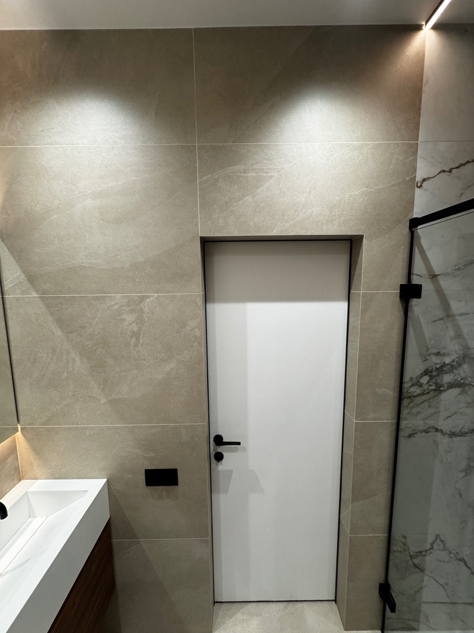 Nowoczesna łazienka z beżowymi płytkami imitującymi kamień, białą umywalką z szafką z drewnianym frontem i białą, minimalistyczną, ukrytą w ścianie, wewnętrzną ościeżnicą z czarną klamką.