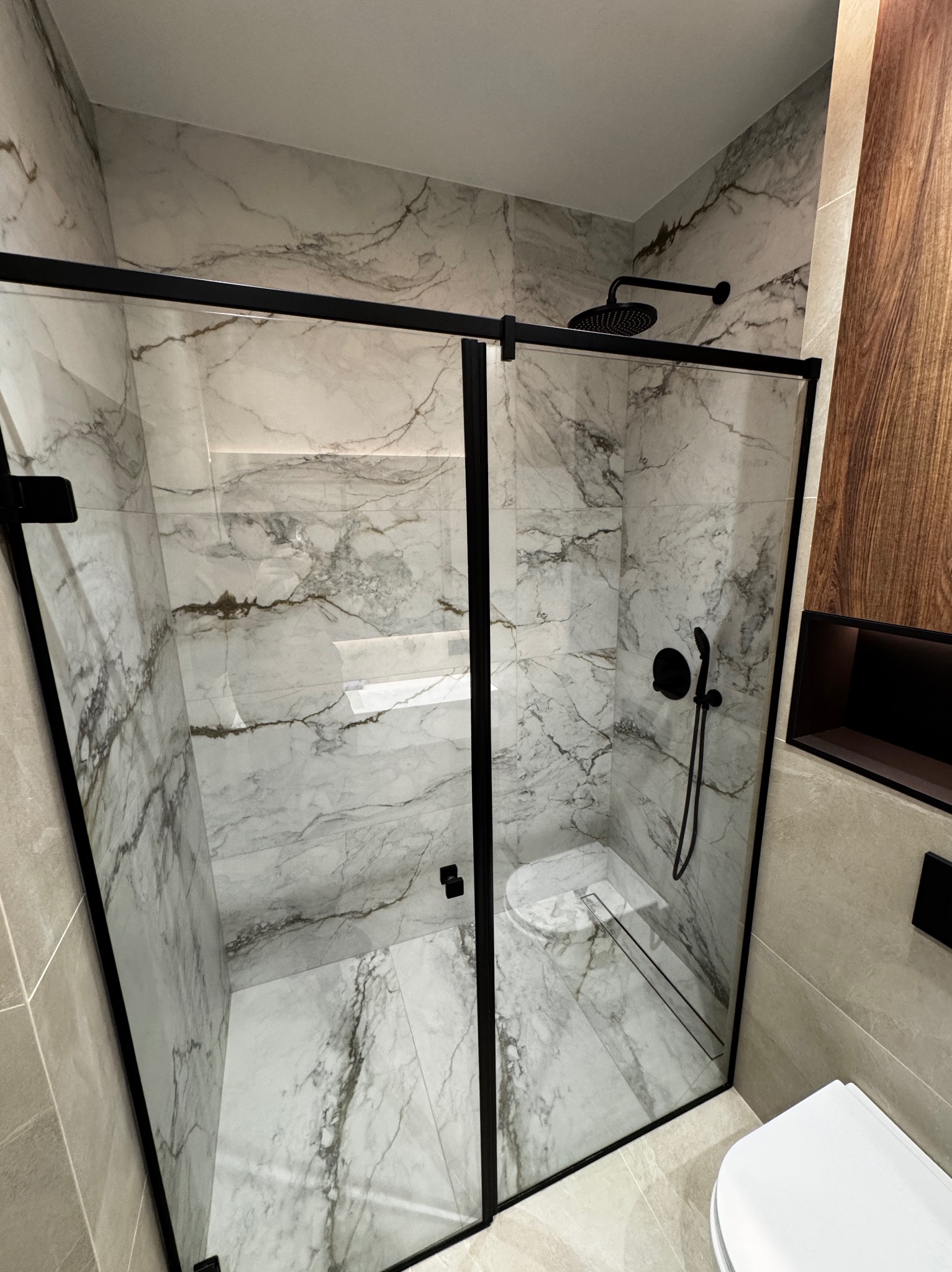 Nowoczesna kabina prysznicowa z czarnymi ramami i deszczownicą, marmurowe płytki na ścianach i podłodze, minimalistyczny design, widoczny fragment toalety.