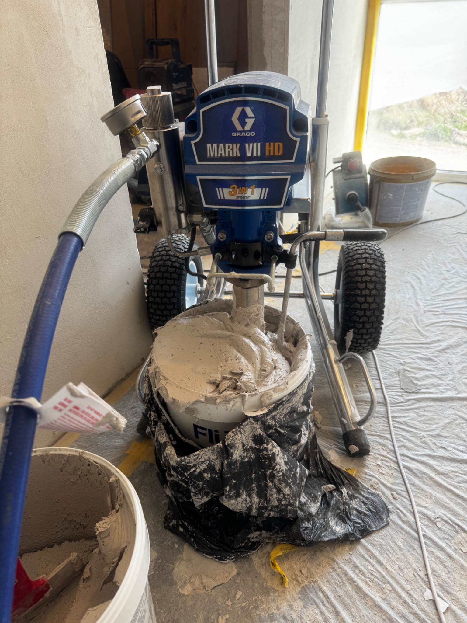 Agregat malarski Graco Mark VII HD z mieszadłem w wiadrze z farbą, przygotowany do pracy w Warszawie. Podłoże zabezpieczone folią malarską.