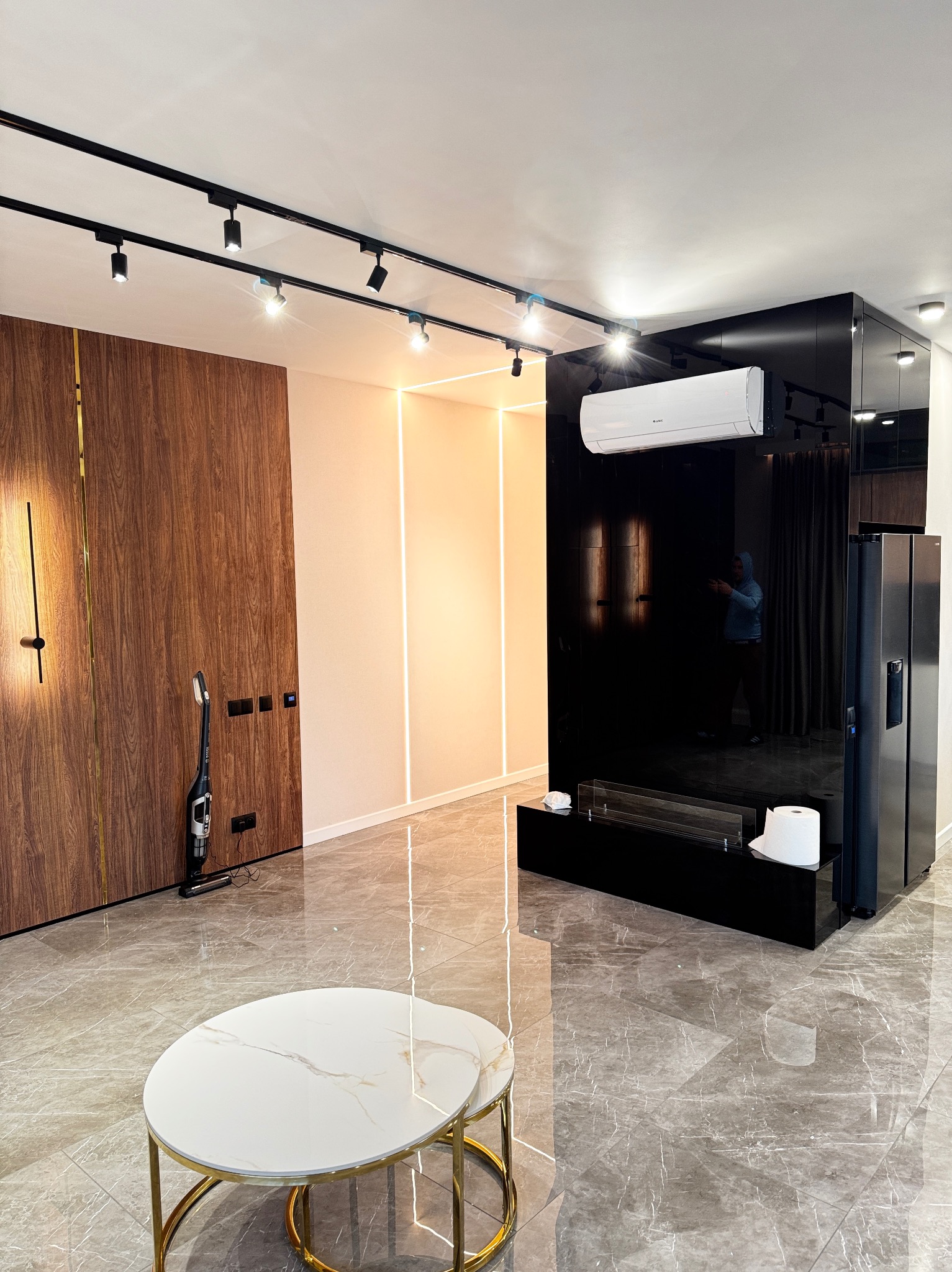 Nowoczesny salon z drewnianą ścianą z pionowym oświetleniem, marmurową podłogą i czarną ścianą z kominkiem oraz klimatyzacją, okrągły stolik kawowy z białym blatem i złotymi nogami na pierwszym...