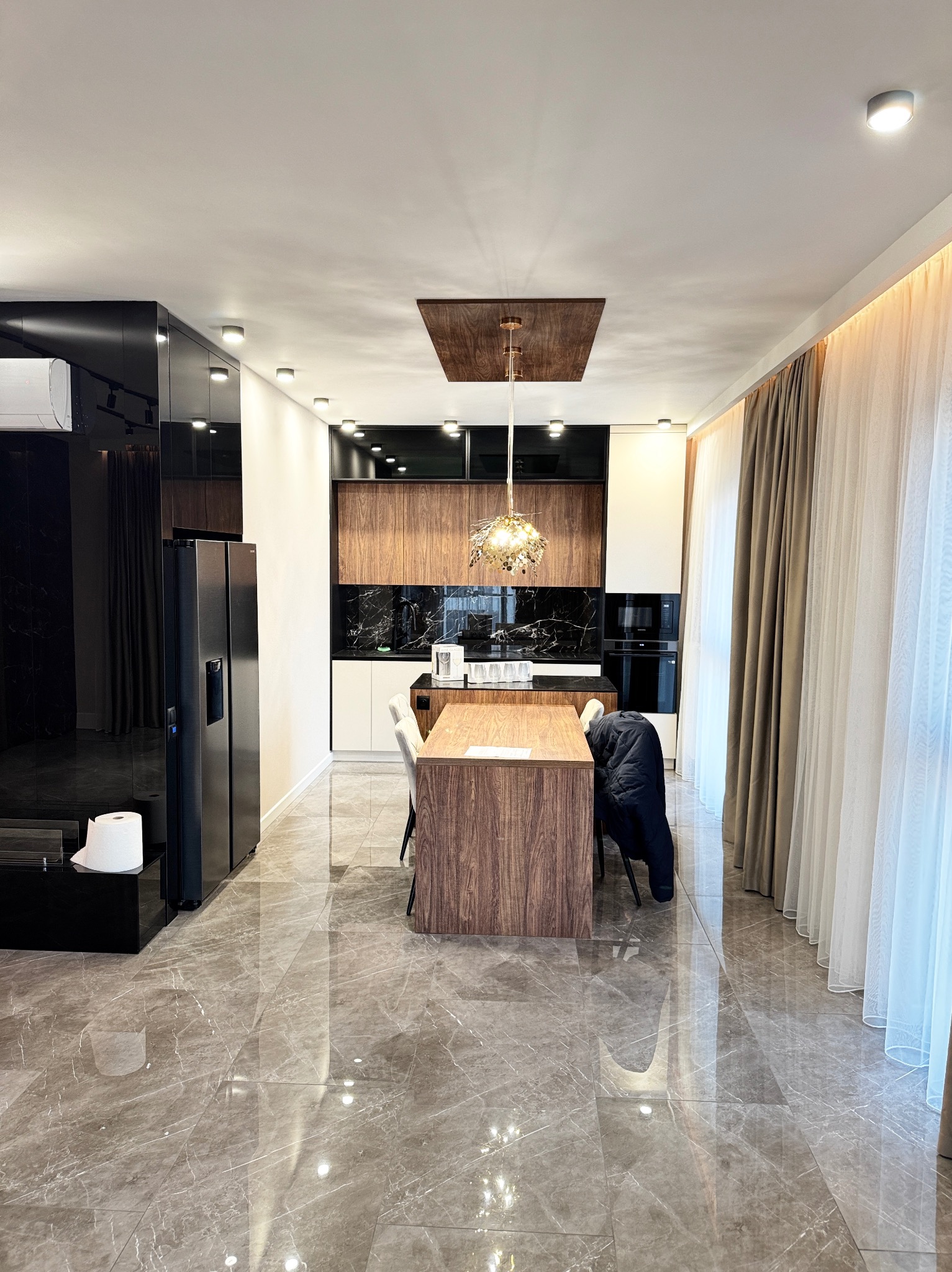 Nowoczesna kuchnia z marmurową podłogą, drewnianymi szafkami i blatem z wyspą, oświetlona dekoracyjną lampą wiszącą, z widocznymi zasłonami w oknie.