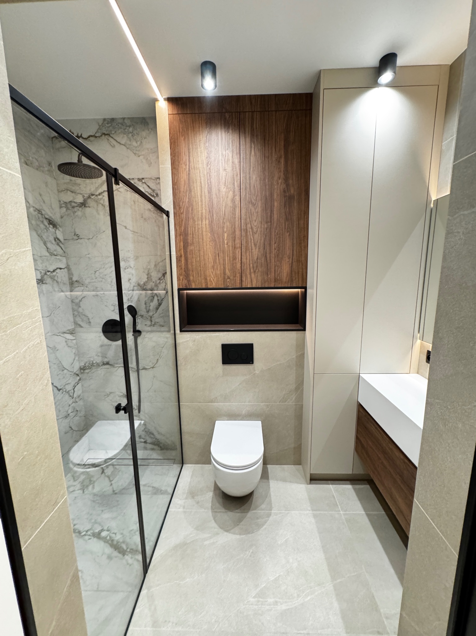 Nowoczesna łazienka z kabiną prysznicową typu walk-in z czarnymi profilami, marmurowymi płytkami na ścianach w kabinie, podwieszaną toaletą i szafką z drewnianym fornirem oraz oświetleniem LED.