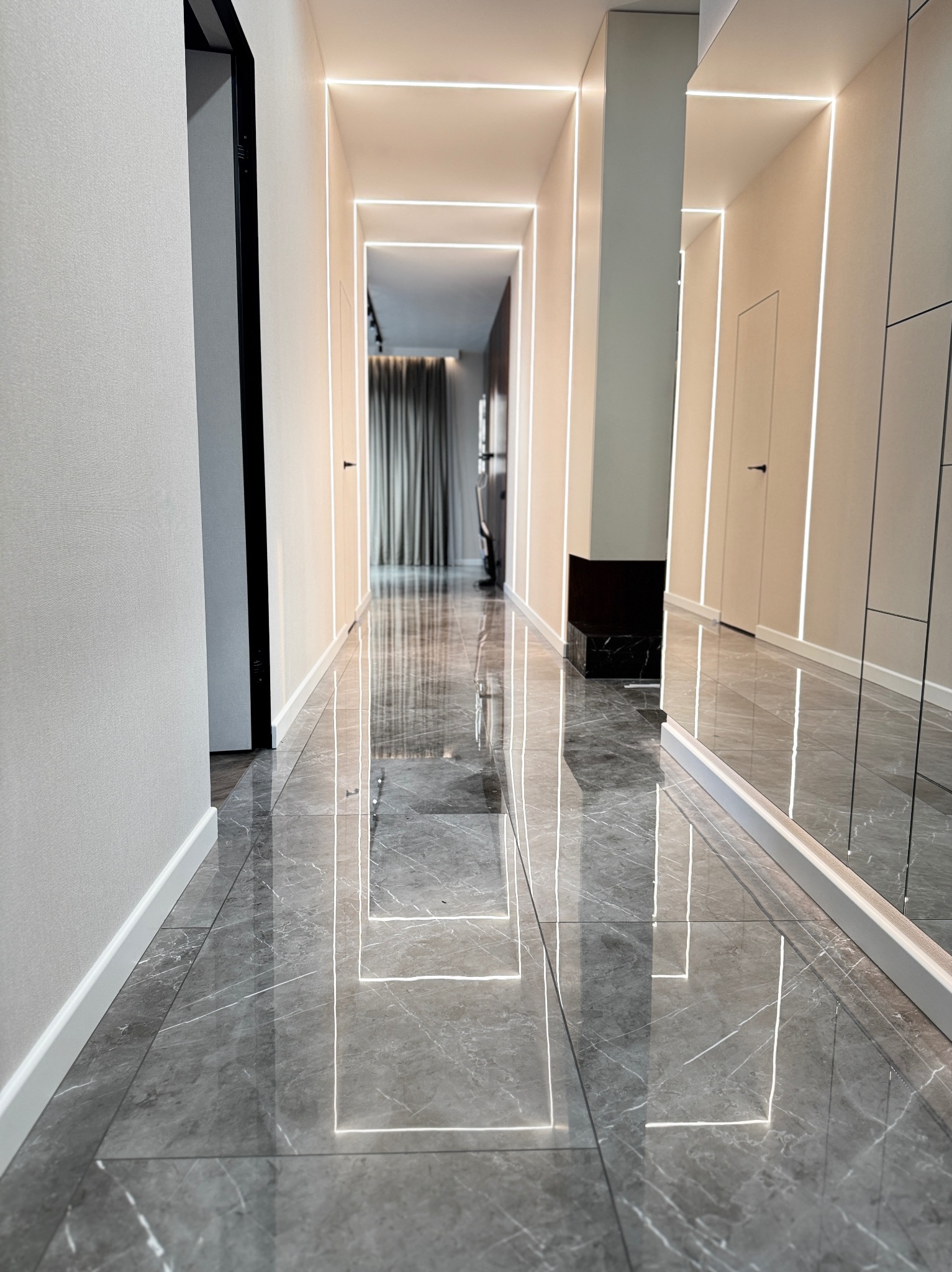 Długi korytarz z szarymi marmurowymi płytkami podłogowymi, oświetleniem LED wzdłuż ścian i sufitu, oraz lustrem na ścianie po prawej stronie.