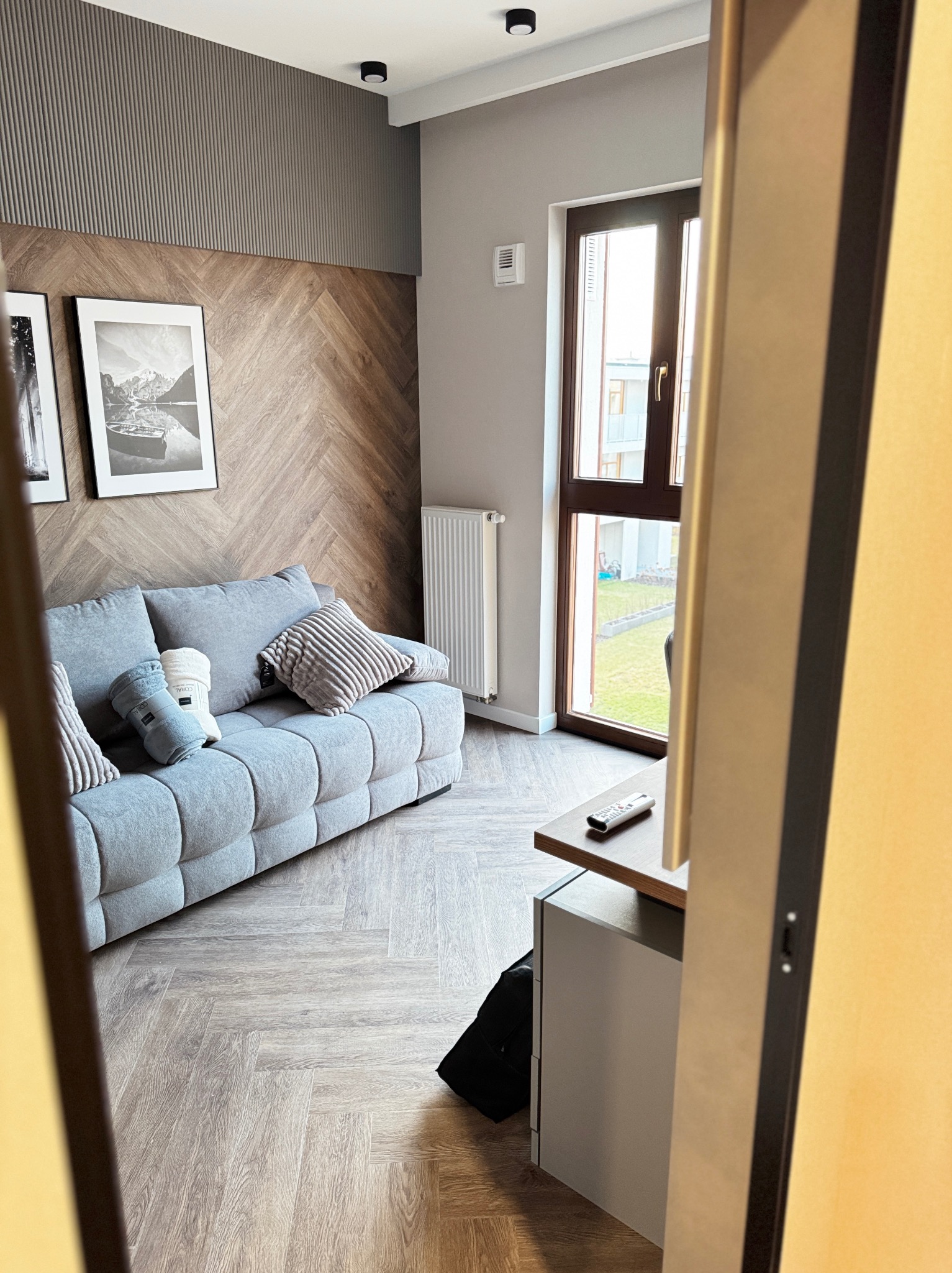 Salon z szarą sofą modułową, drewnianą ścianą w jodełkę, obrazami i widokiem na zewnątrz przez okno, widziany zza framugi drzwi.