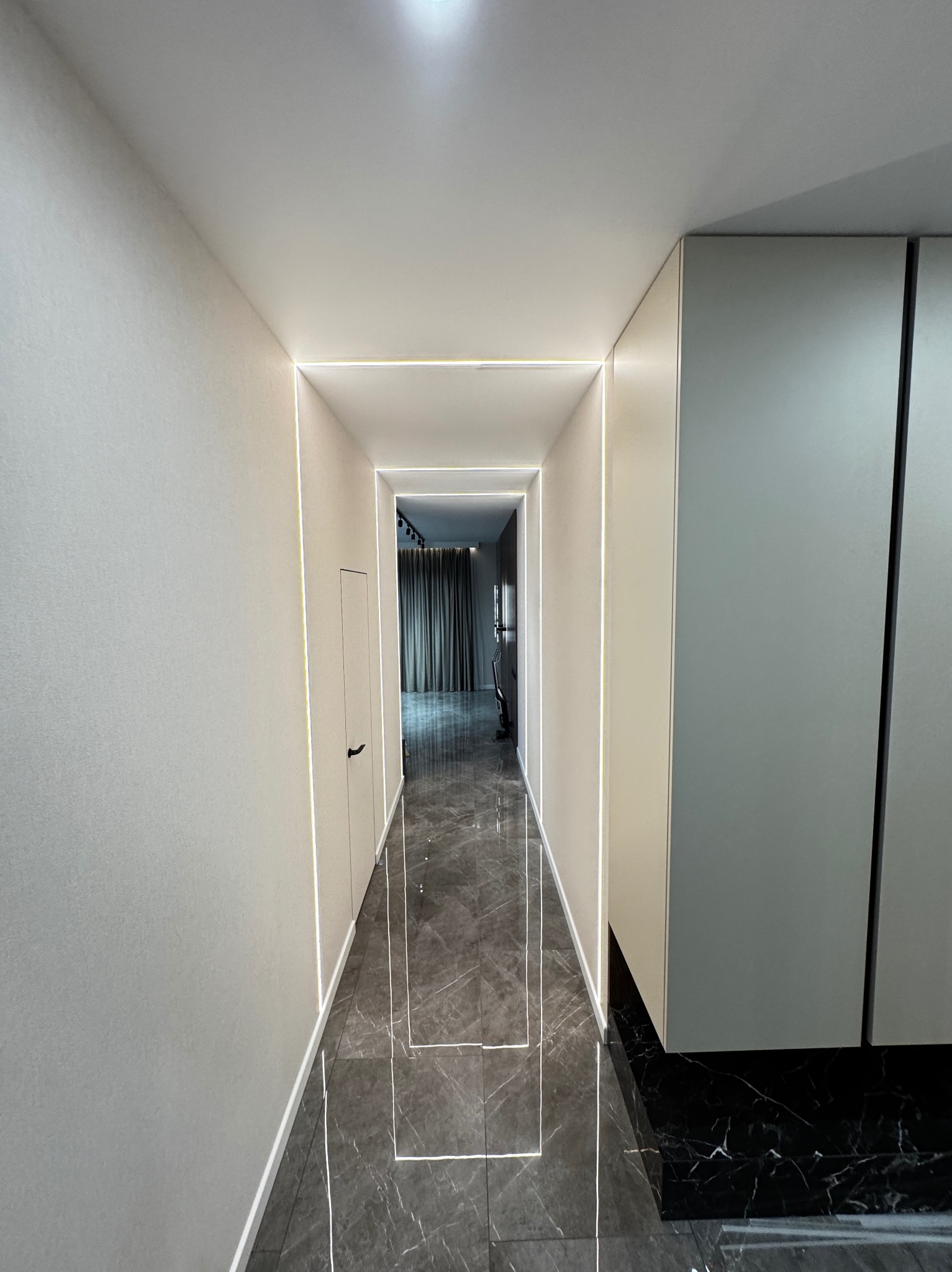 Korytarz z szarymi ścianami, podłogą z płytek imitujących marmur i oświetleniem LED wzdłuż krawędzi ścian i podłogi, perspektywa z głębi korytarza.