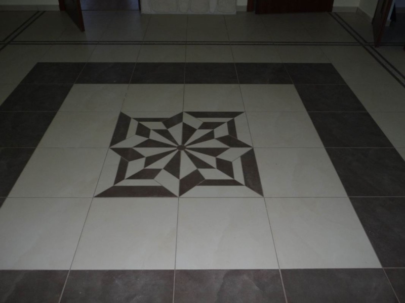 Wzór geometryczny z płytek podłogowych w odcieniach beżu i brązu, ułożony w kwadratową kompozycję centralną, z widocznymi fugami i fragmentami drzwi w tle.