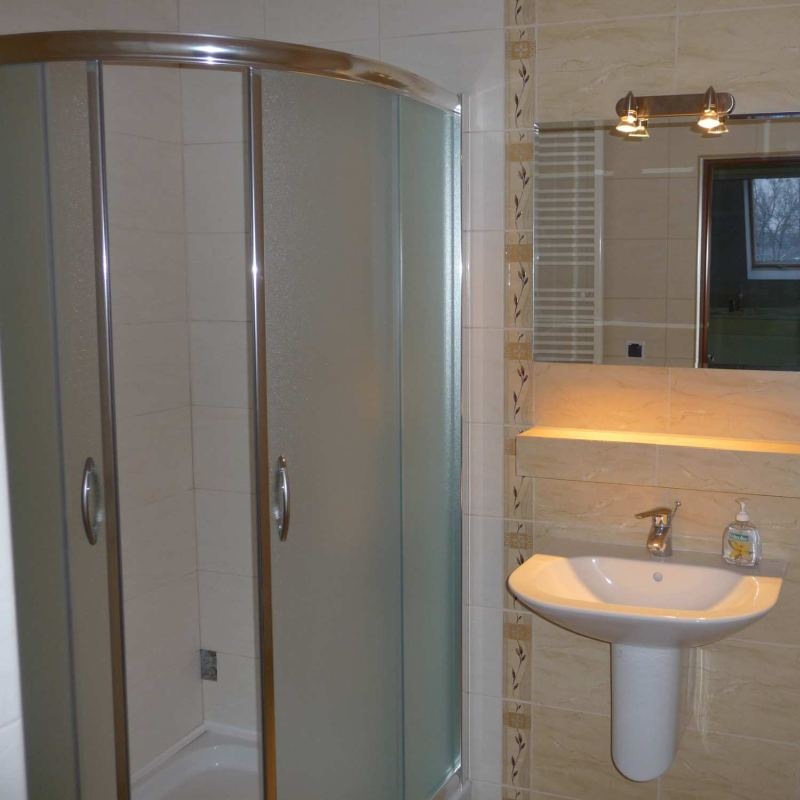 Wykończona łazienka z kabiną prysznicową z przesuwnymi drzwiami, umywalką na postumencie i beżowymi płytkami z dekoracyjnymi wstawkami.