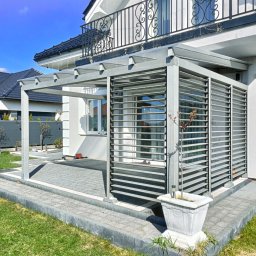CleanWood - Nowoczesna, szara pergola z poziomymi lamelami przy białym domu z balkonem, obok kostka brukowa i trawnik